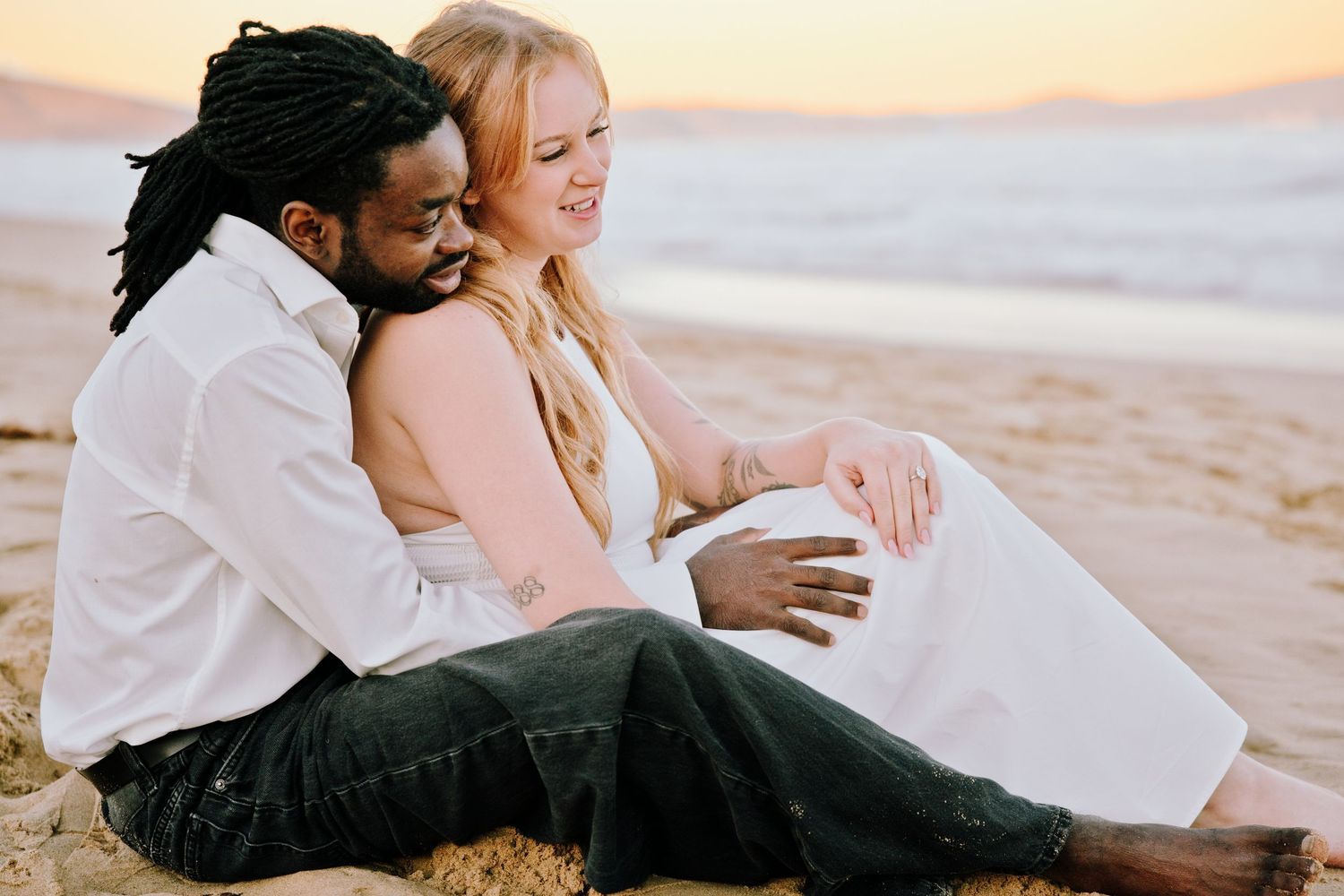 Interracial couple sitting on beach at sunset, woman in white dress leaning against man in white shirt, smiling together.