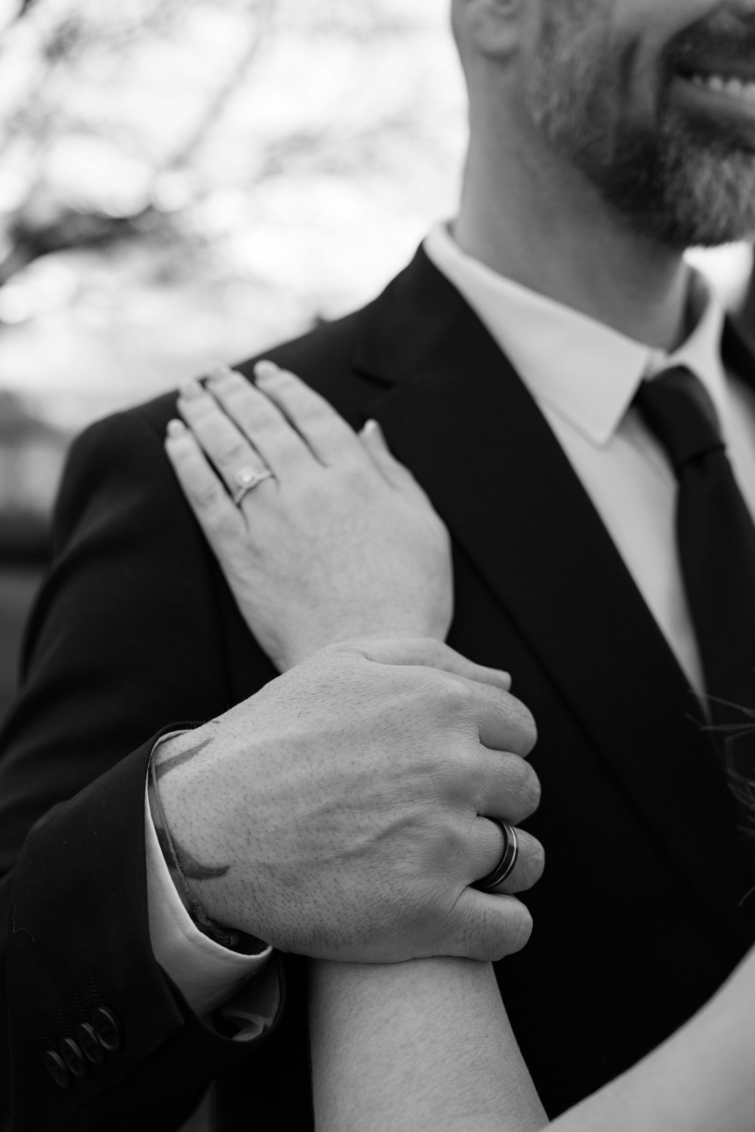 Black and white photo of couple holding hands, showing wedding rings, groom in suit and tie smiling.