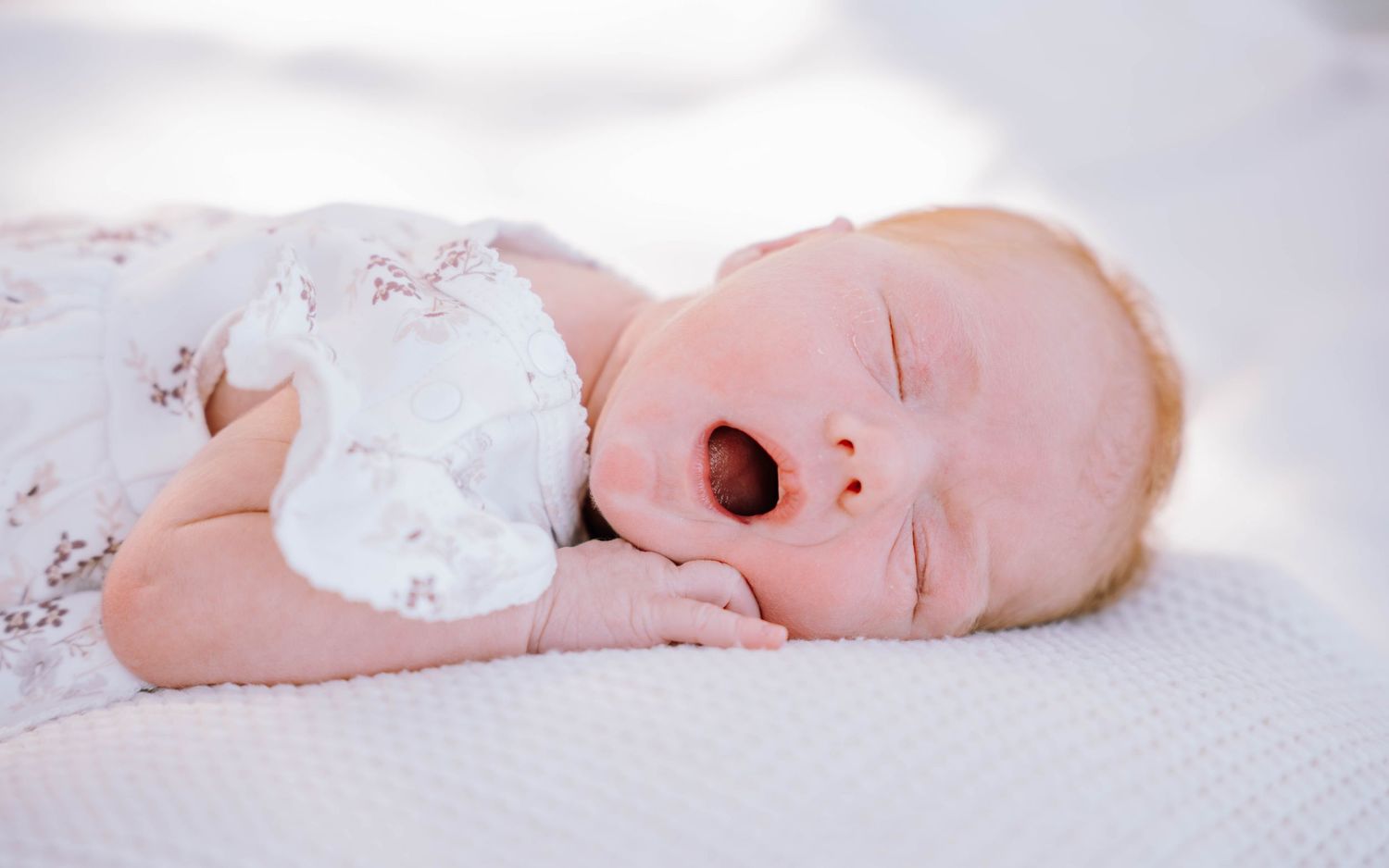 Newborn baby yawning while sleeping peacefully on a white pillow, dressed in a floral onesie.