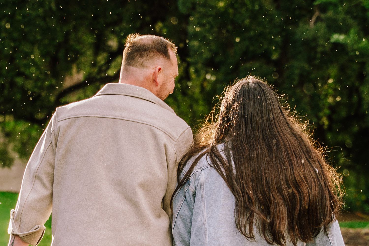 Couple walking together outdoors near green trees, viewed from behind, man in beige coat and woman with long brown hair.