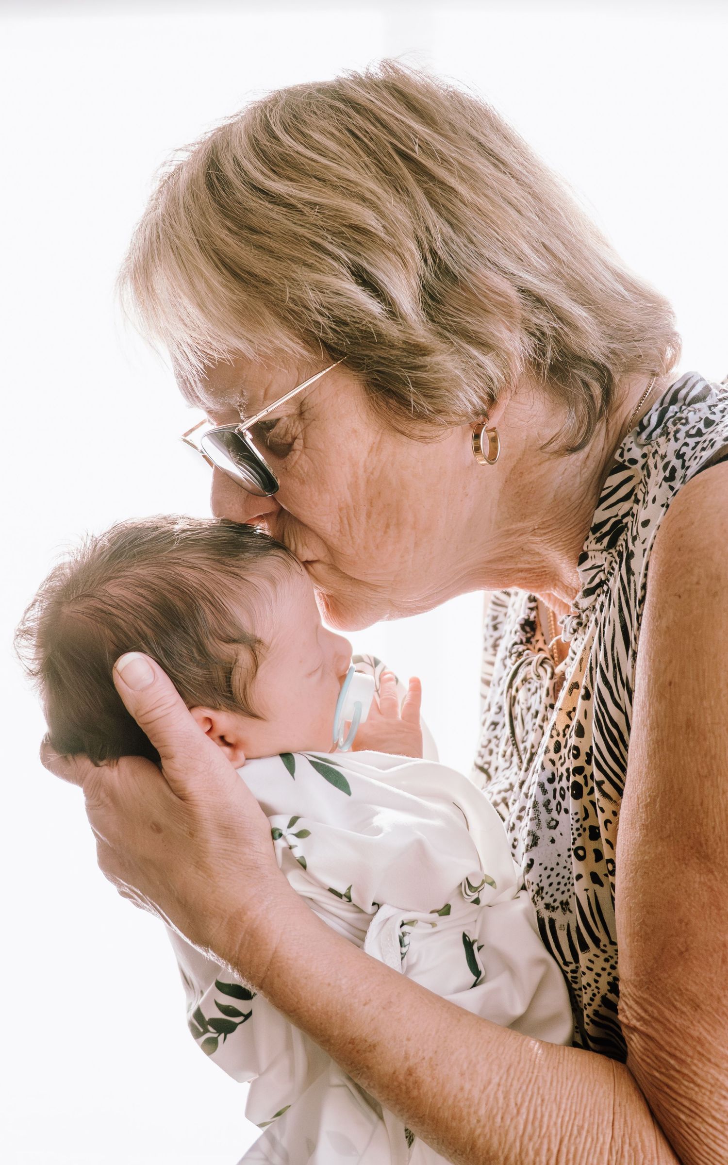 A tender moment captured as a woman in glasses and floral dress gently kisses a baby's head.