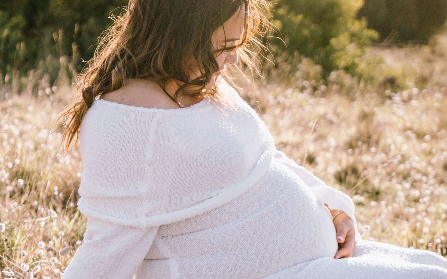 A person in a white sweater sits in a sunlit field of dried grass and wildflowers during golden hour.