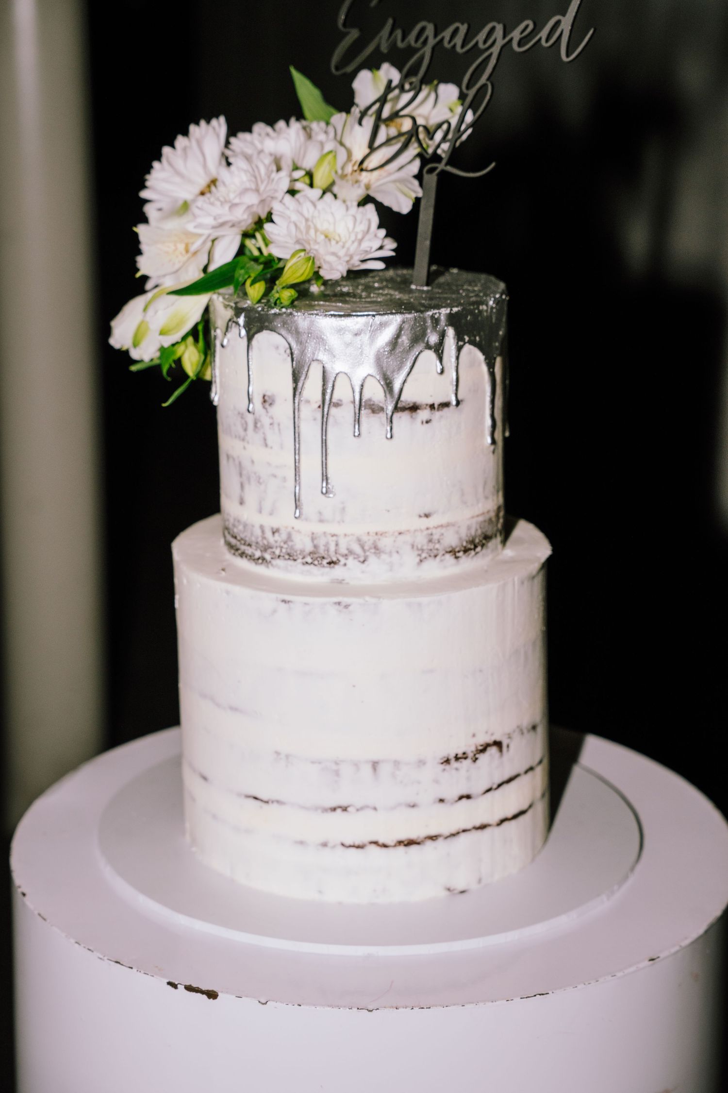 Two-tier naked engagement cake with silver drip topping and white floral arrangement, displayed on white pedestal stand.