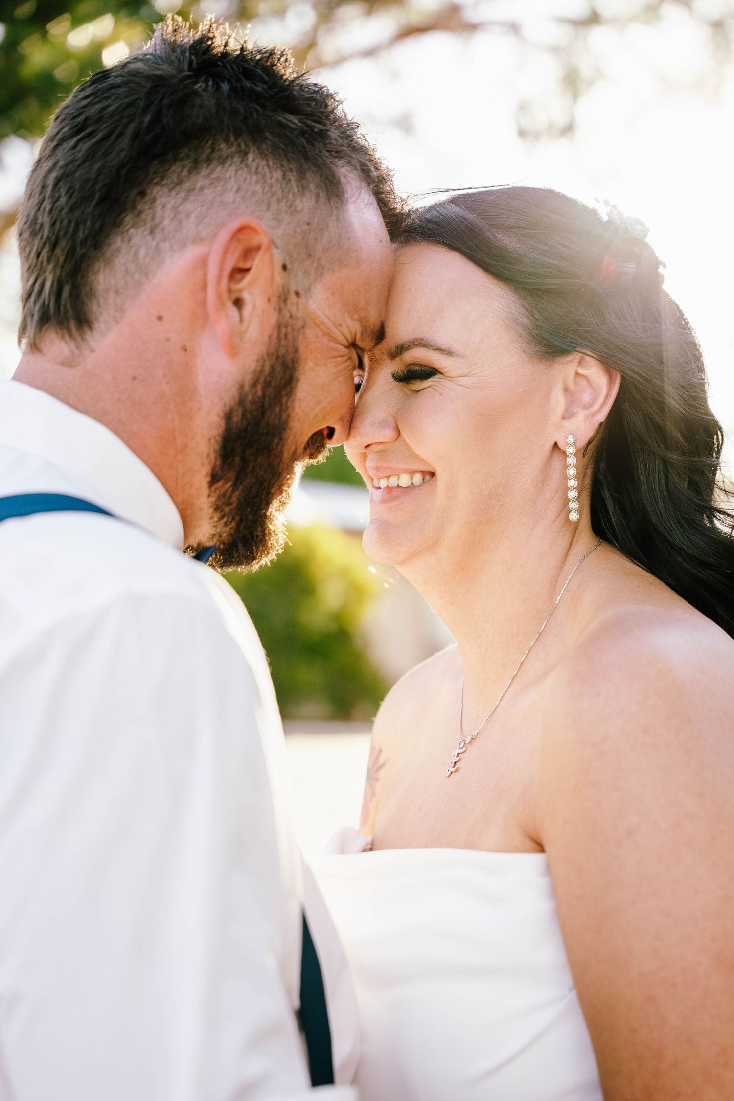 Bride and groom touching foreheads and smiling on their wedding day outdoors in warm sunlight.