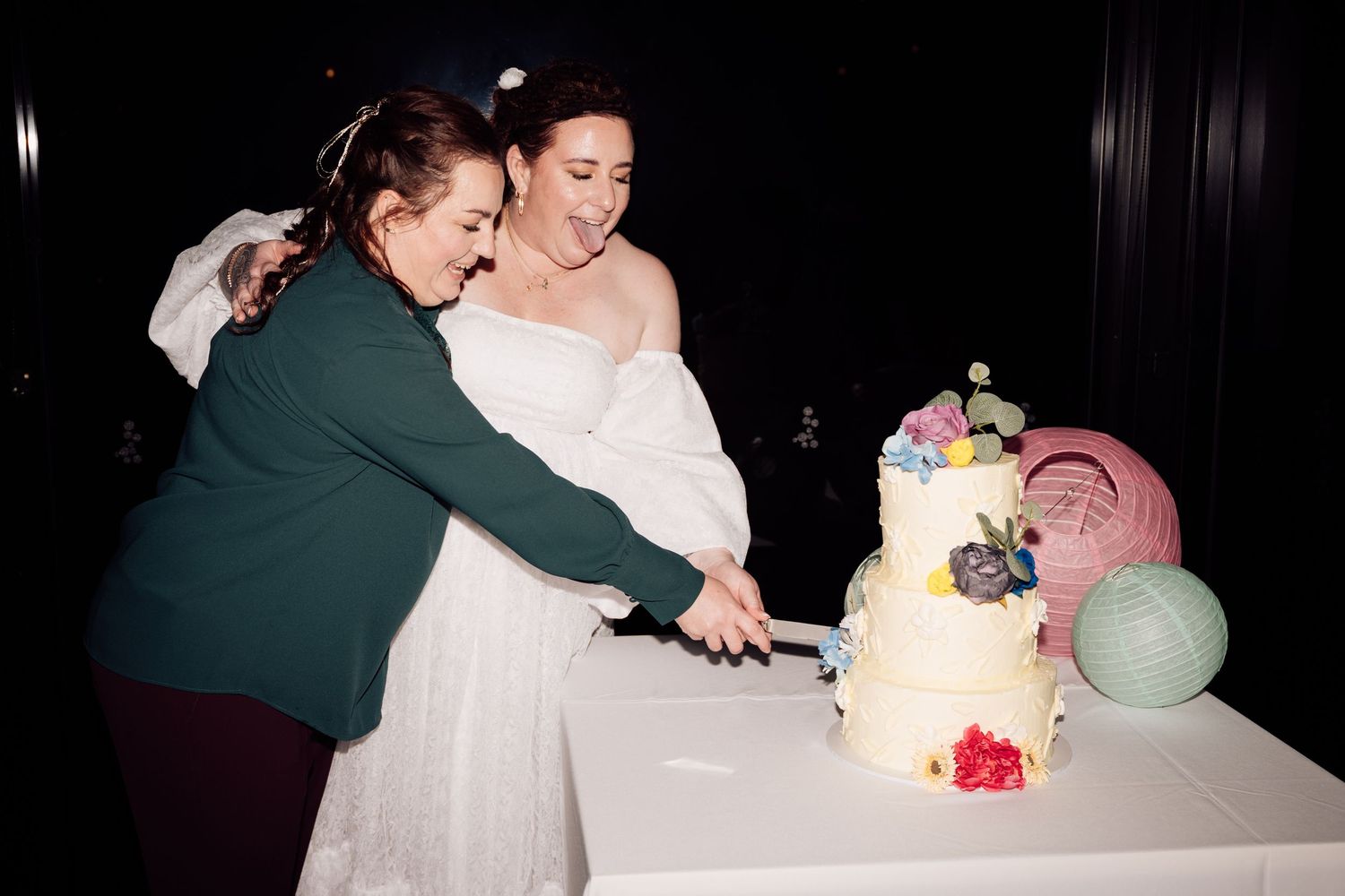 Two women cutting a decorated three-tier wedding cake with colorful flowers at a wedding reception.