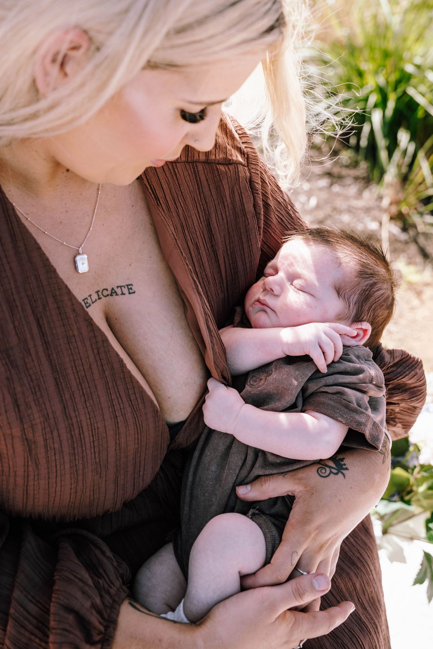 Blonde mother tenderly cradling her sleeping newborn baby dressed in brown outdoors.