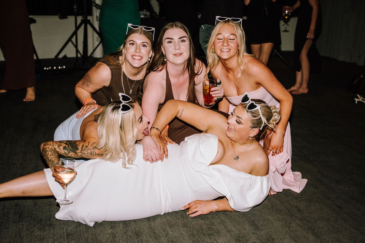 Group of five women posing together at a party, wearing sunglasses and pink outfits, smiling on a dance floor.