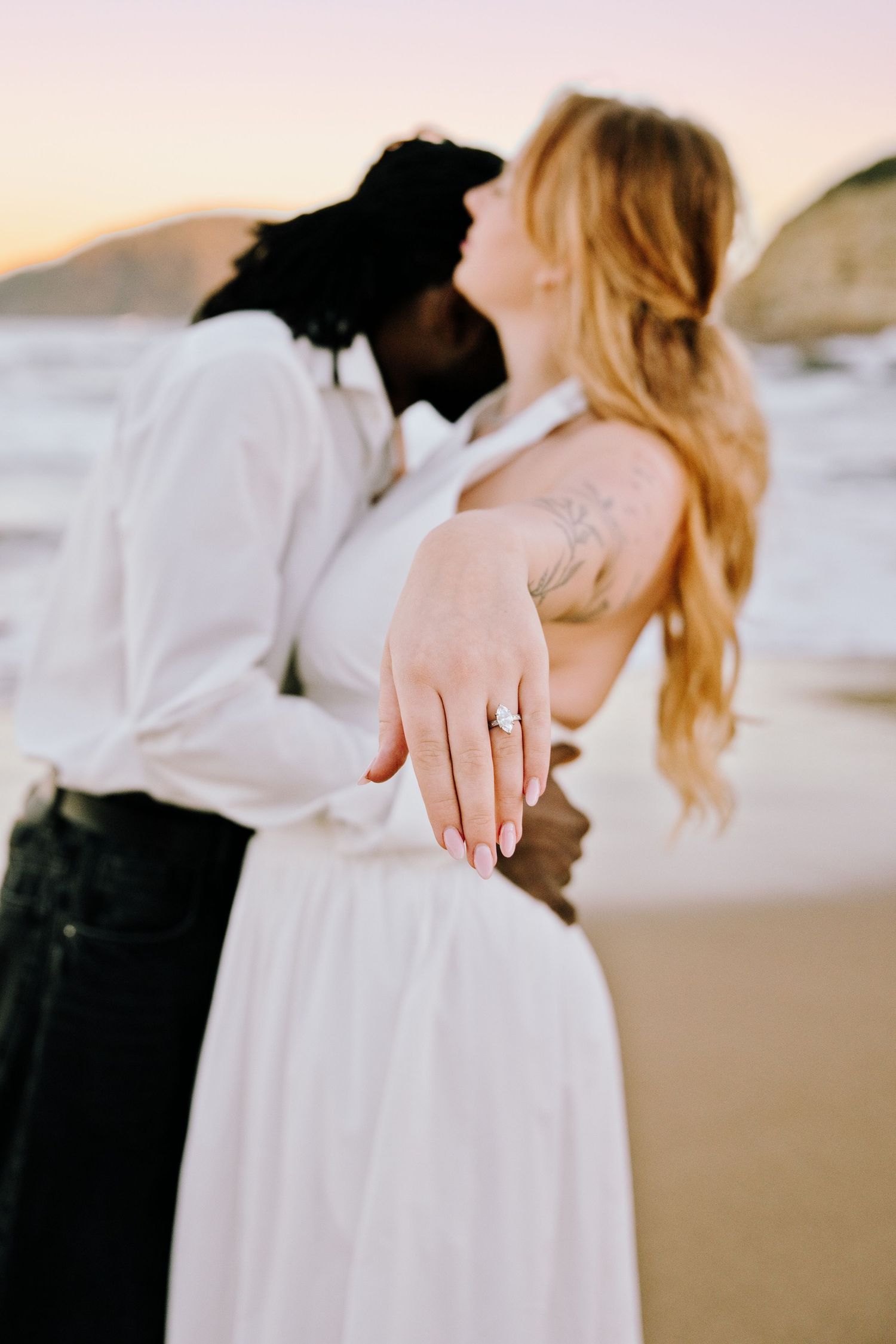 Interracial couple kissing on beach at sunset, woman showing engagement ring, wearing white outfits with ocean backdrop.