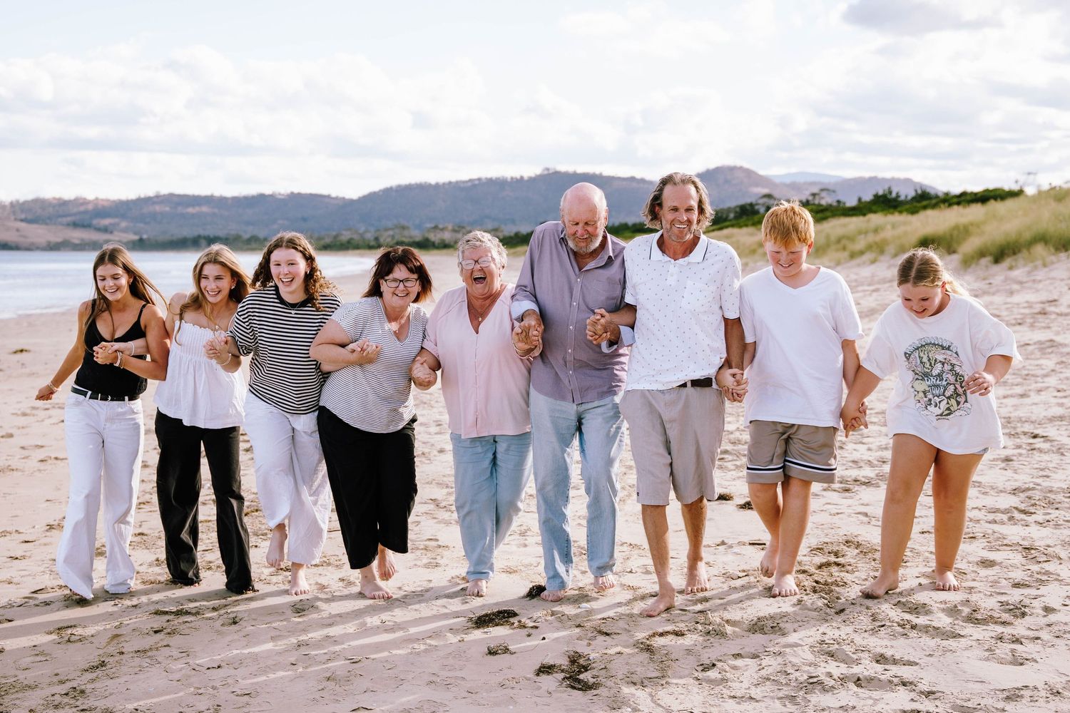 A joyful multigenerational family laughing and walking together on a sandy beach with scenic hills in the background.