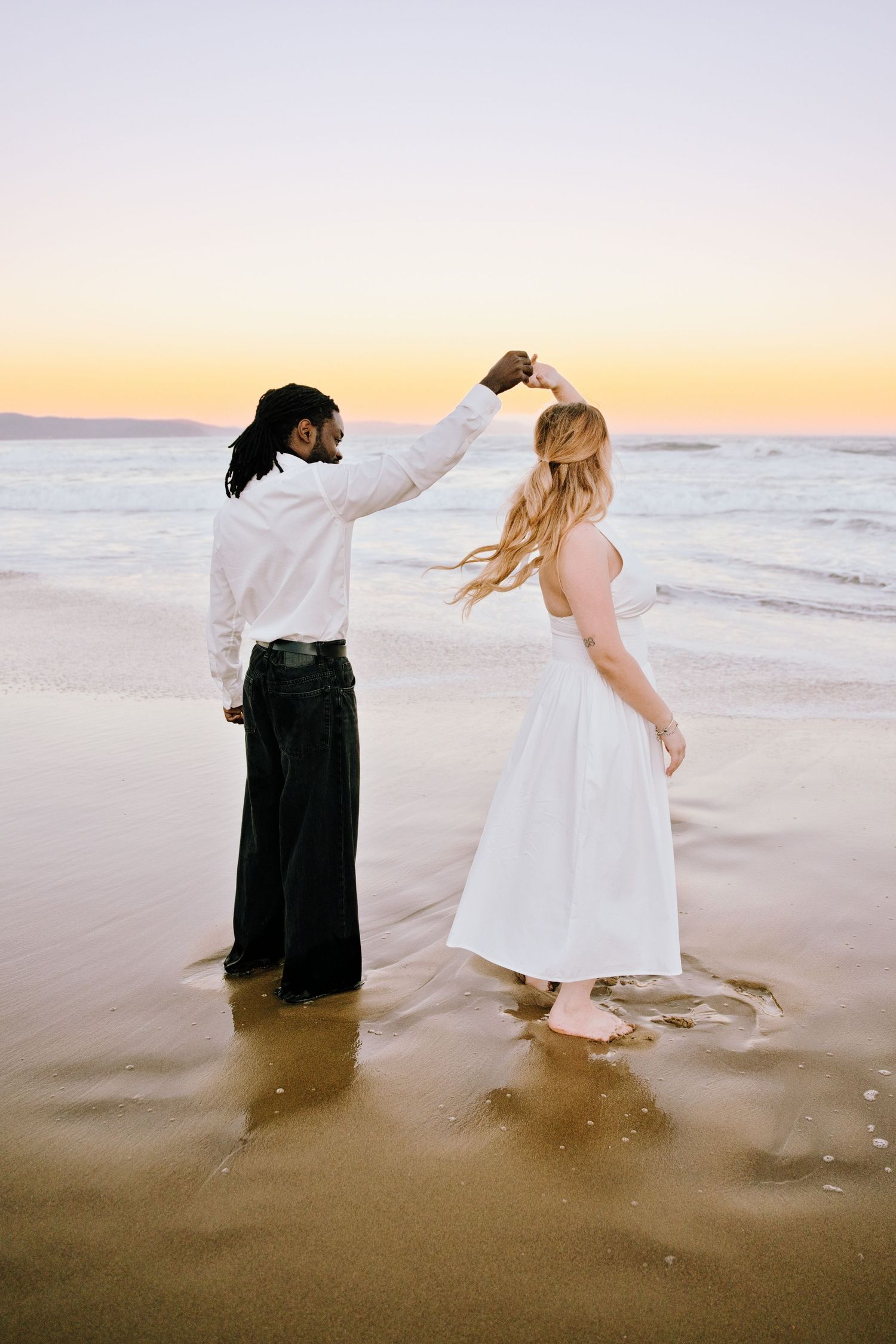 Couple dancing on beach at sunset, man in white shirt spinning woman in white dress near ocean waves.