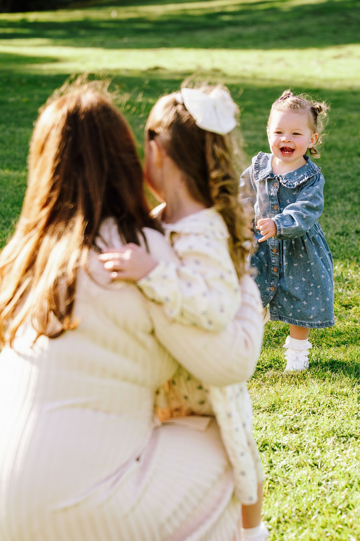 A mother and older daughter kneel on grass as a laughing toddler girl in denim dress runs toward them.