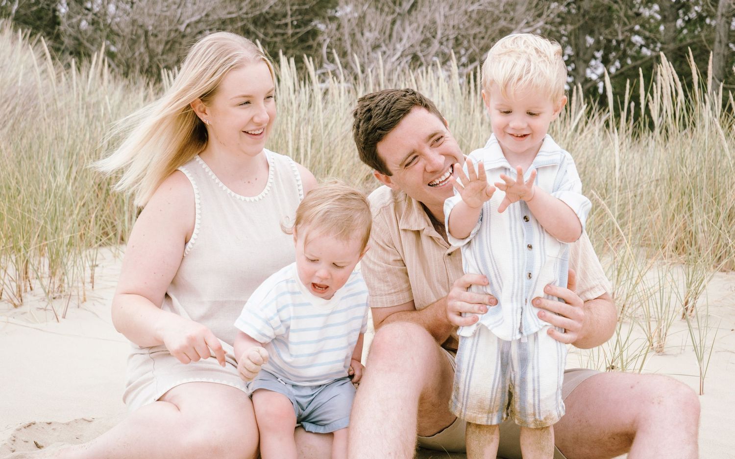 Happy family of four smiling on sandy beach dunes, parents with two young toddler boys on a sunny day.
