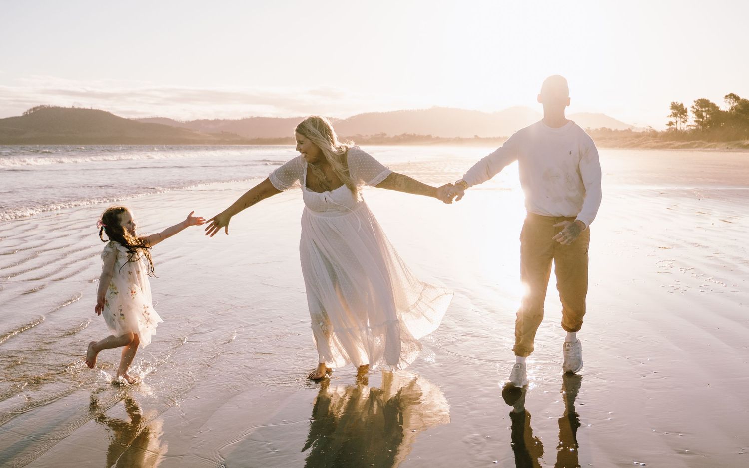A couple walking hand in hand along a beach at sunset while their dogs follow behind them.