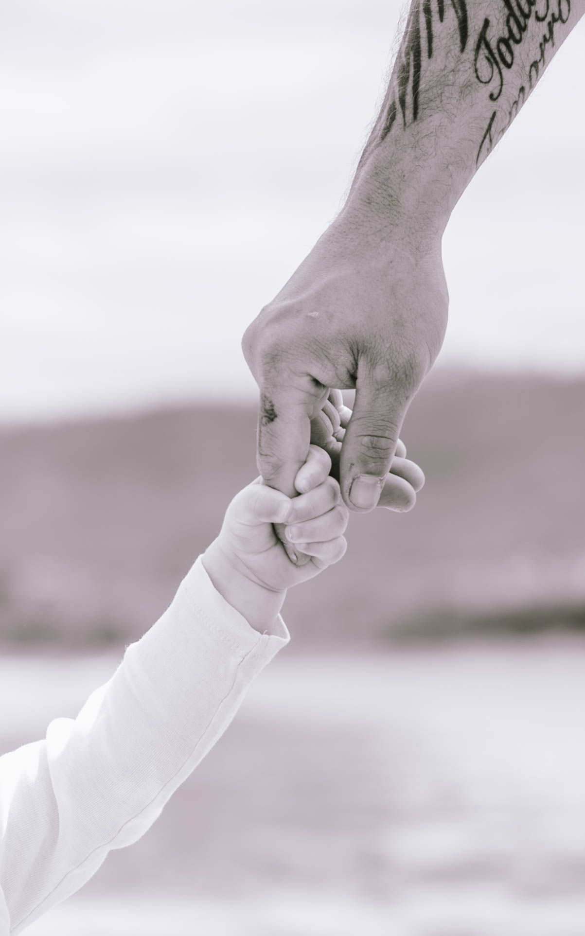 A close-up black and white photo of two hands reaching towards each other, with one appearing to be a child's hand.