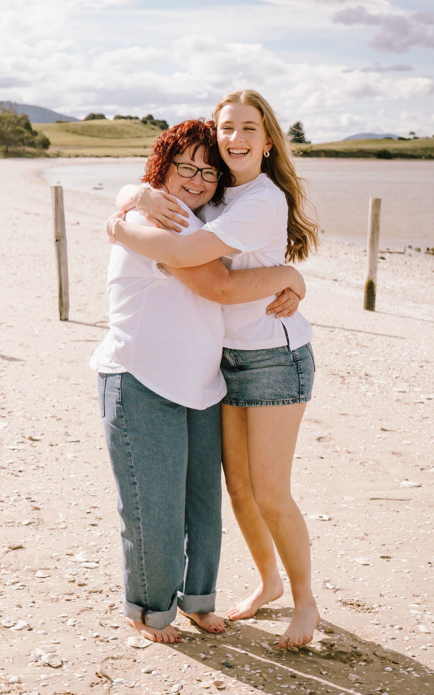 Two people share warm embraces on a sunny beach with rolling hills in the background.