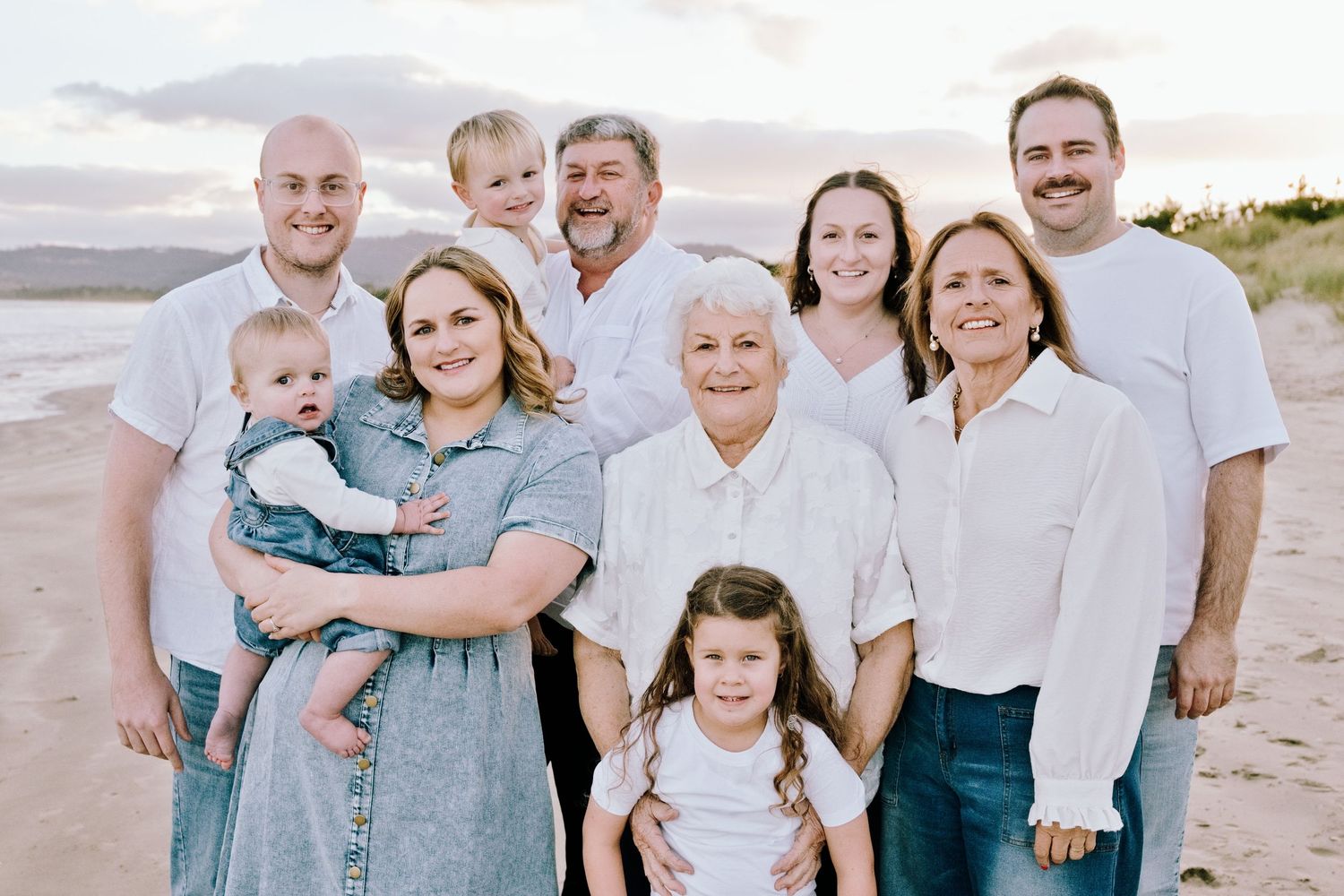 Large family group portrait on a sandy beach at sunset, wearing coordinated white tops and jeans.