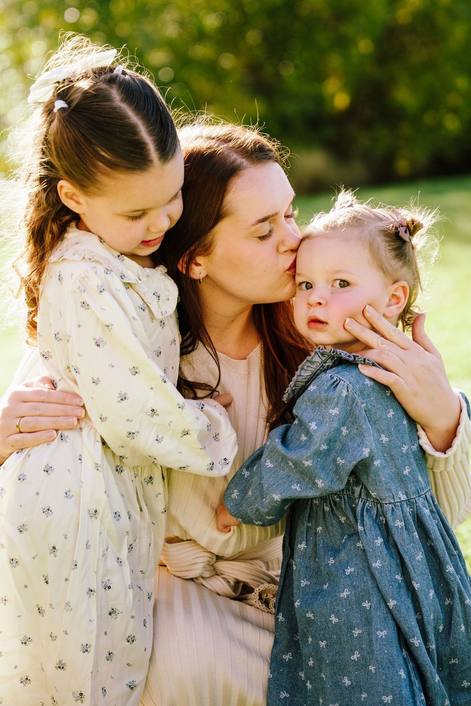 A mother tenderly kisses her toddler's cheek while embracing two young daughters in floral dresses outdoors.