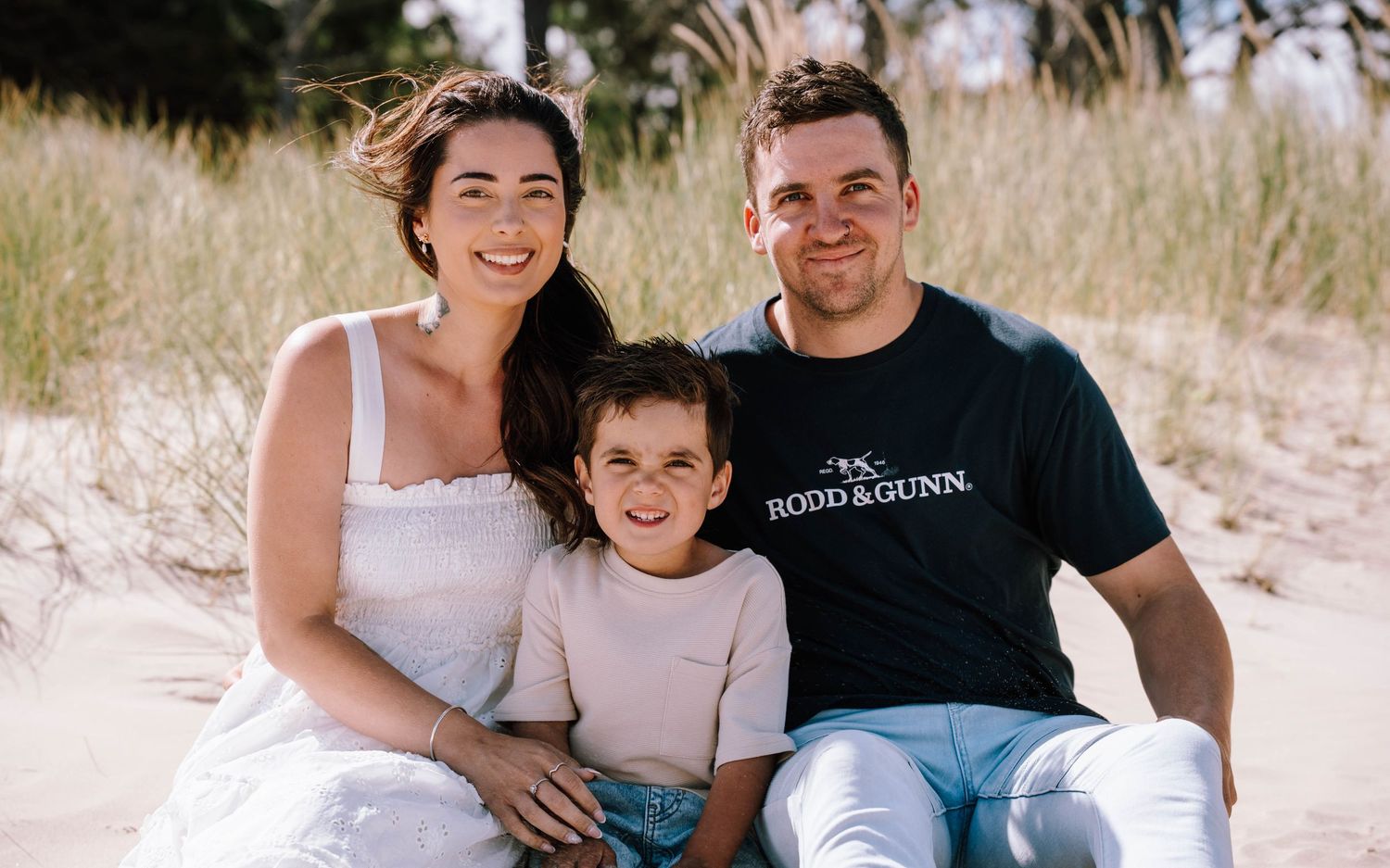 A family group poses together on a sandy beach during a sunny day, wearing casual beach attire.