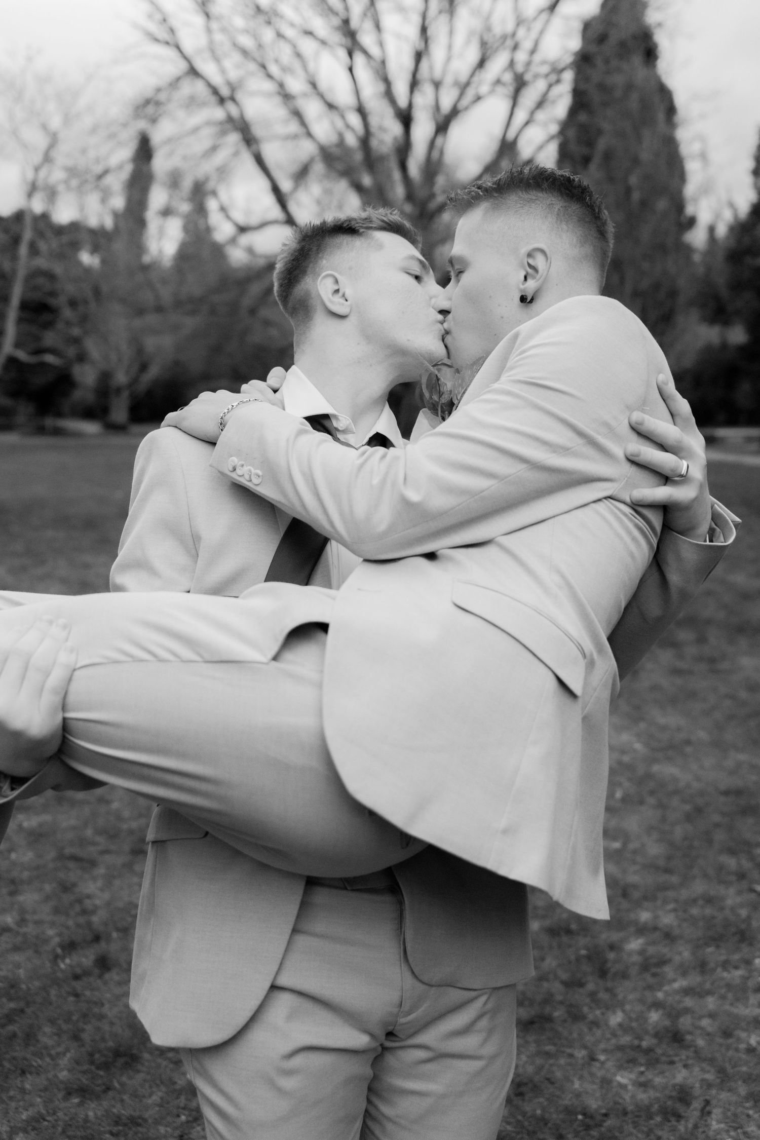 Two grooms share a romantic kiss as one is lifted in a black and white wedding photo.