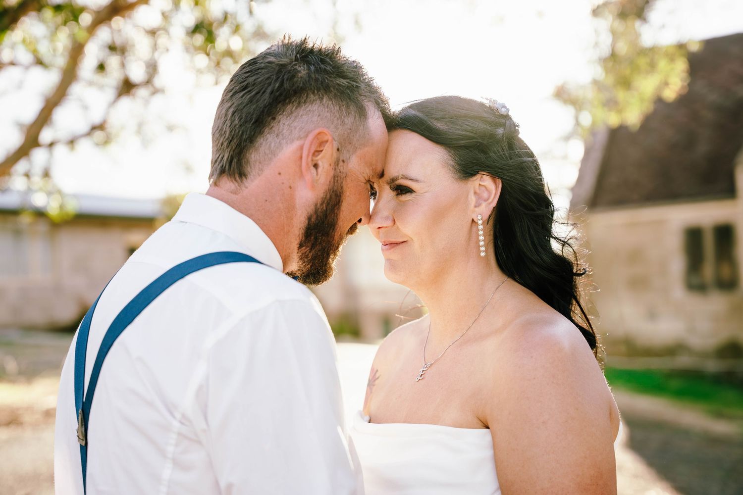 Bride and groom share an intimate forehead touch moment outdoors on their wedding day, groom wearing suspenders.