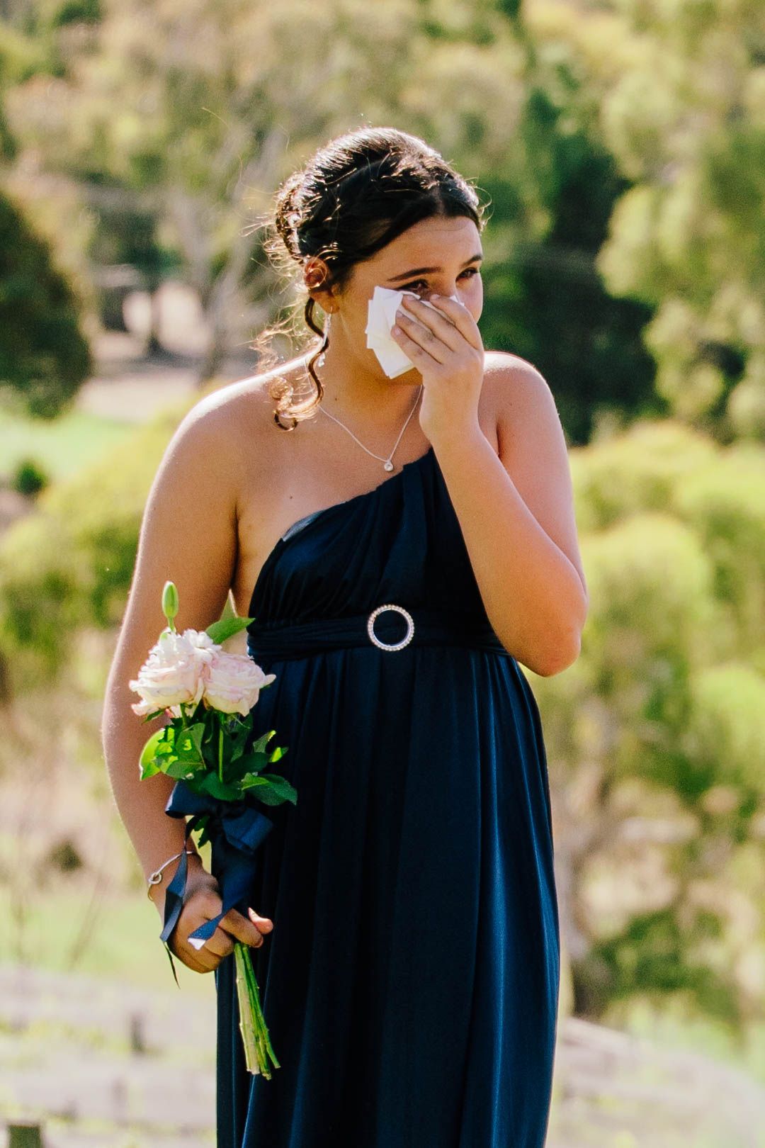Emotional bridesmaid in navy blue dress crying while holding white roses bouquet at outdoor wedding ceremony.