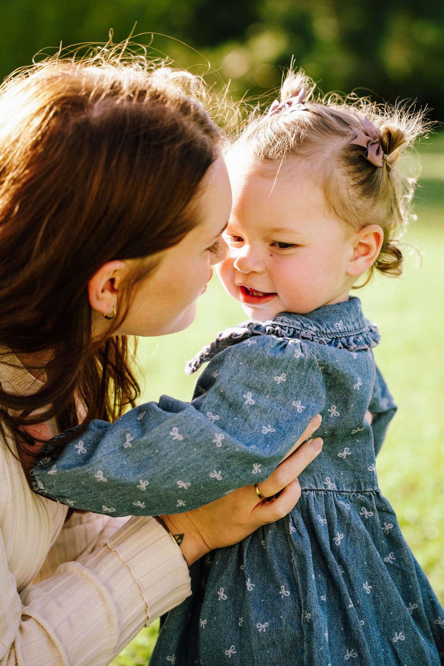 A mother tenderly holds her smiling toddler girl in a sunny green park, both sharing a joyful moment.