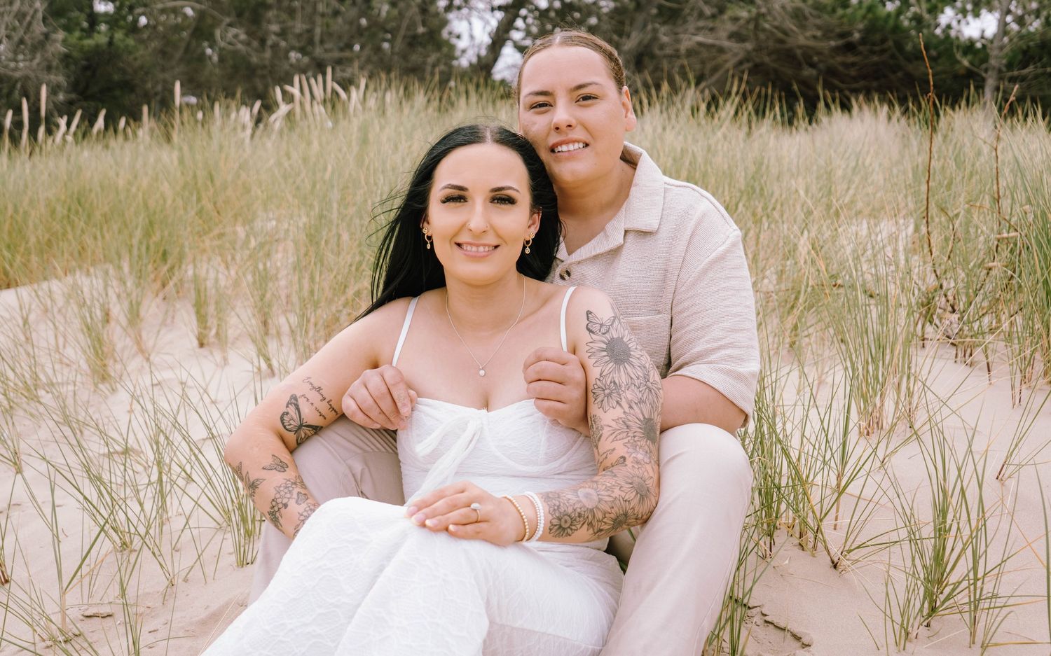 Couple sitting close together on sand dunes surrounded by coastal grass and natural scenery.