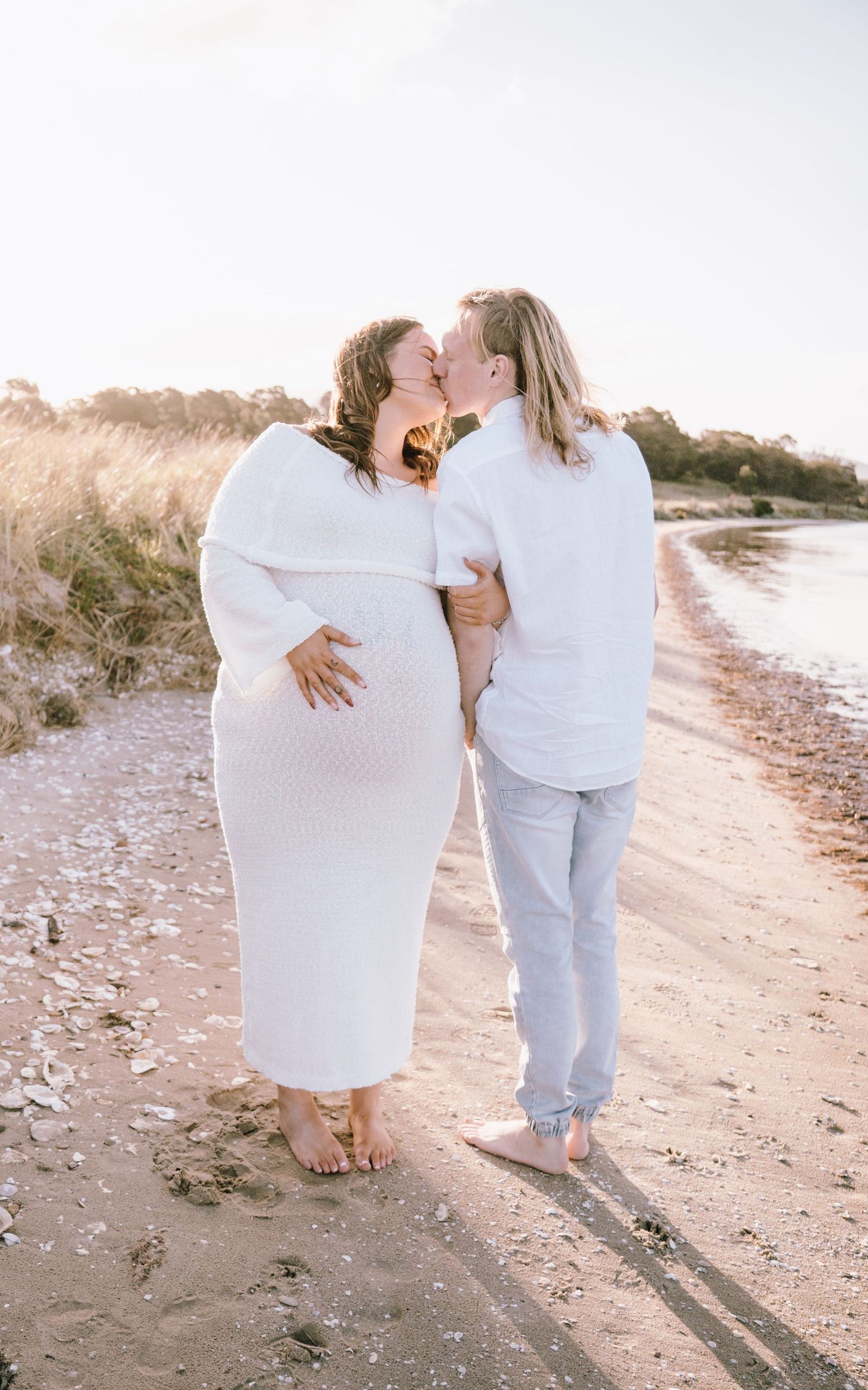 Two people in white outfits walking along a beach shoreline at sunset casting long shadows.