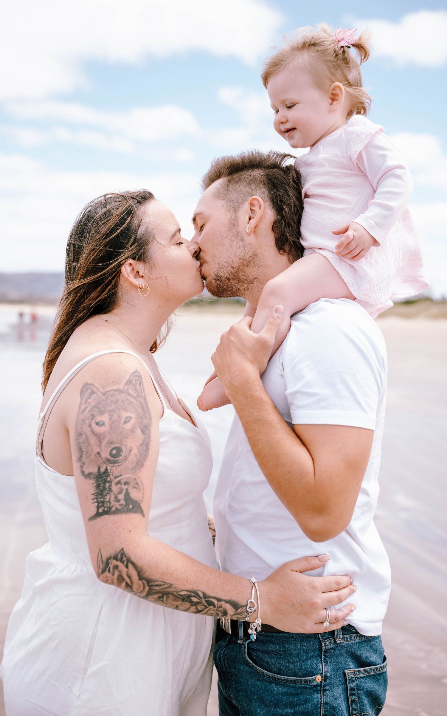 A tender family moment at the beach with a small child in pink perched on shoulders while parents share an intimate kiss.
