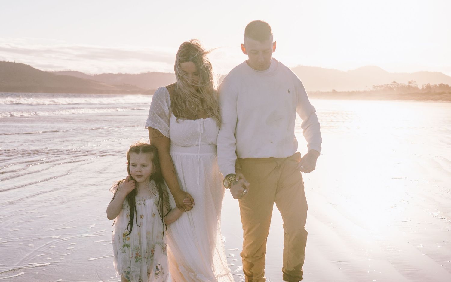 A couple walks along a sandy beach at sunset holding hands as waves roll in along the shore.