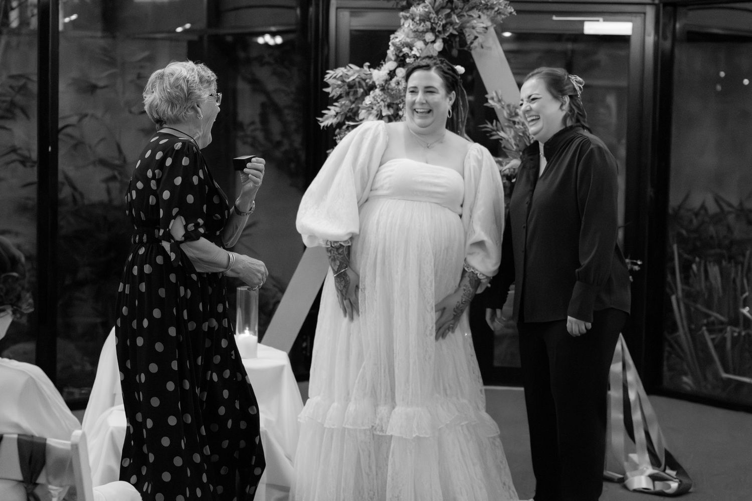 Black and white photo of a pregnant bride in a white gown smiling with two women at a wedding ceremony.