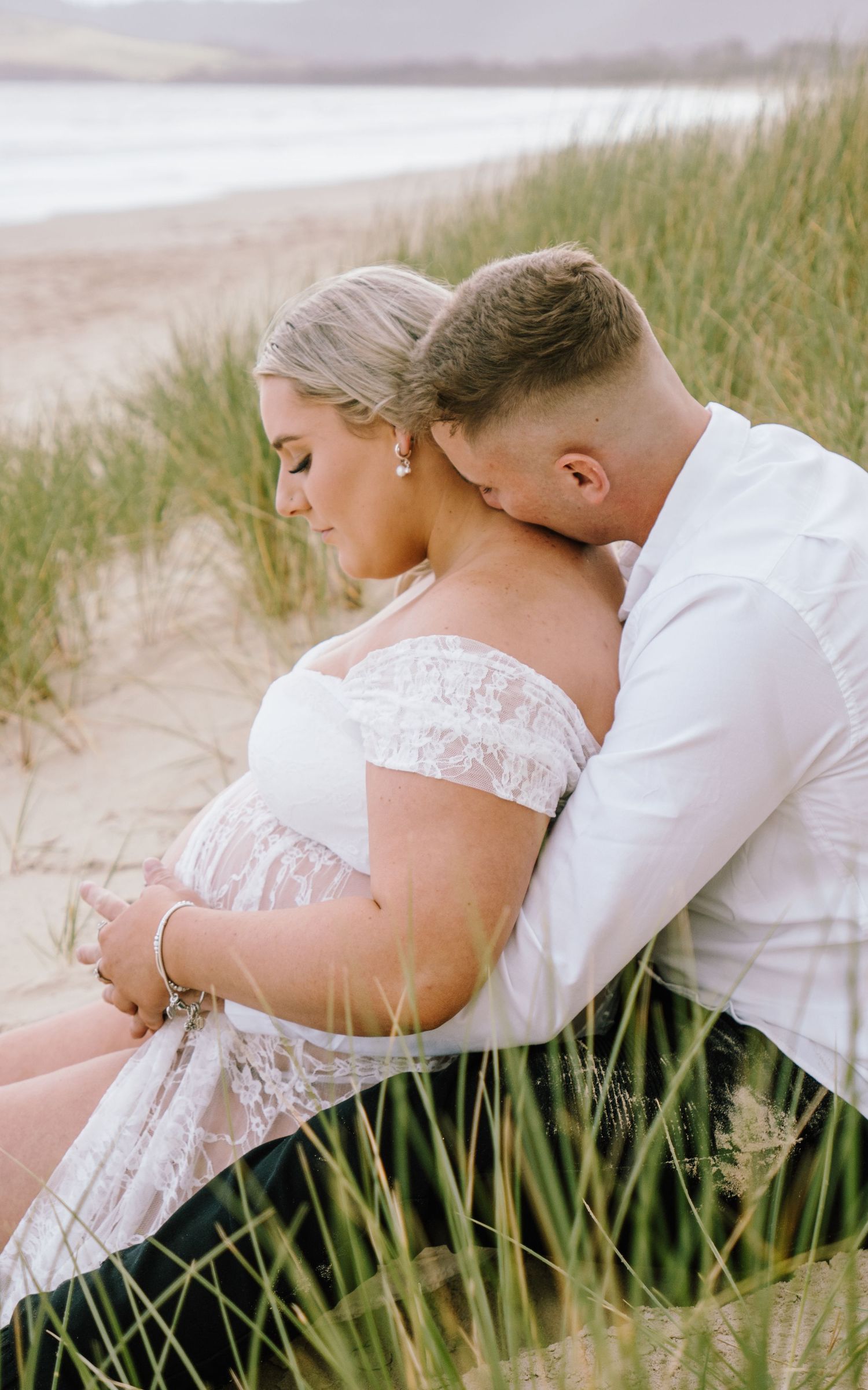 A romantic maternity photo shoot on a beach with tall grass, featuring a couple in white embracing on the sand.