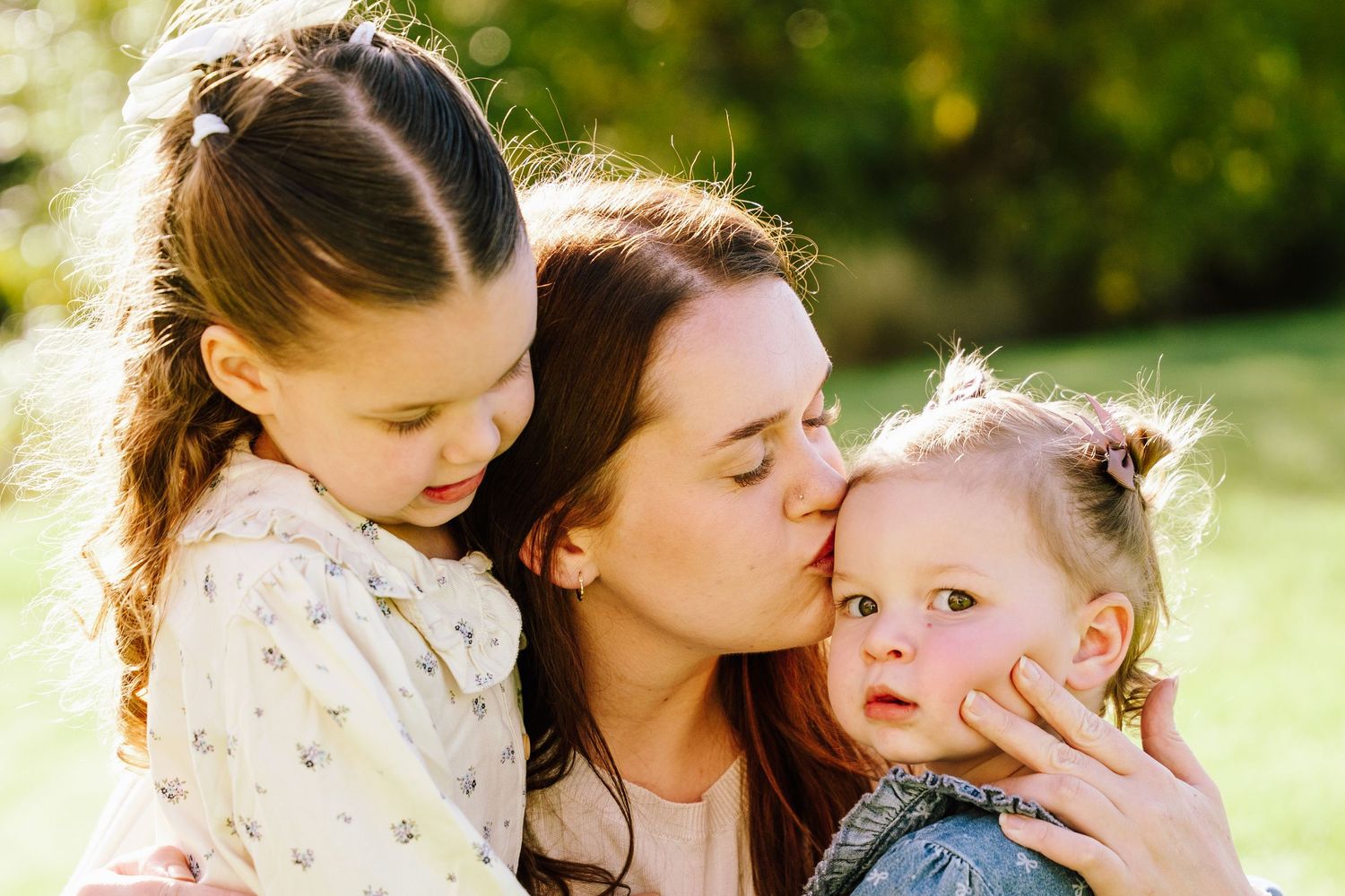A mother tenderly kisses her toddler while embracing her older daughter outdoors in a sunlit green park.