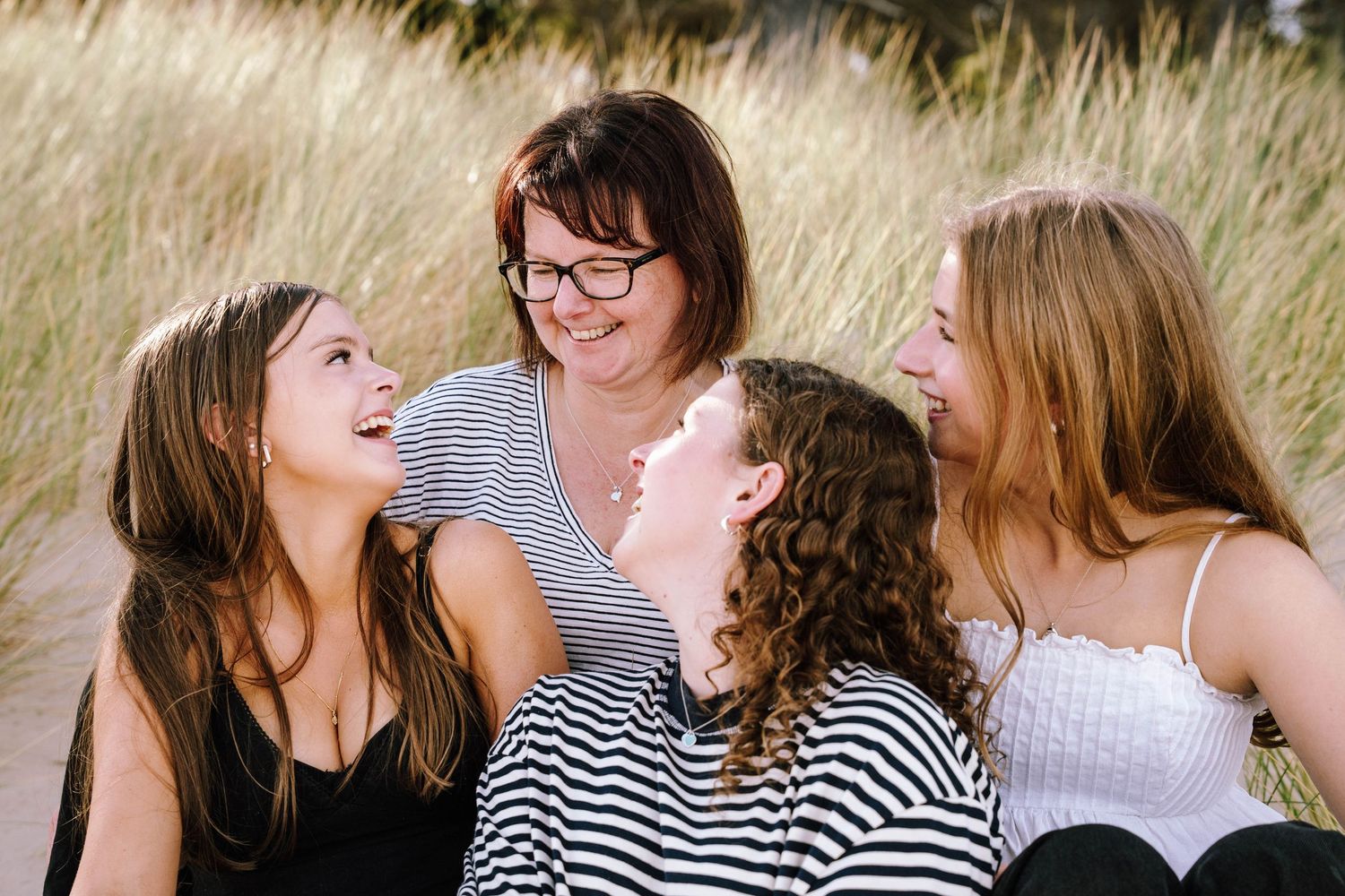 A woman with glasses and three teenage girls laugh together outdoors near sandy dunes in a candid family portrait.