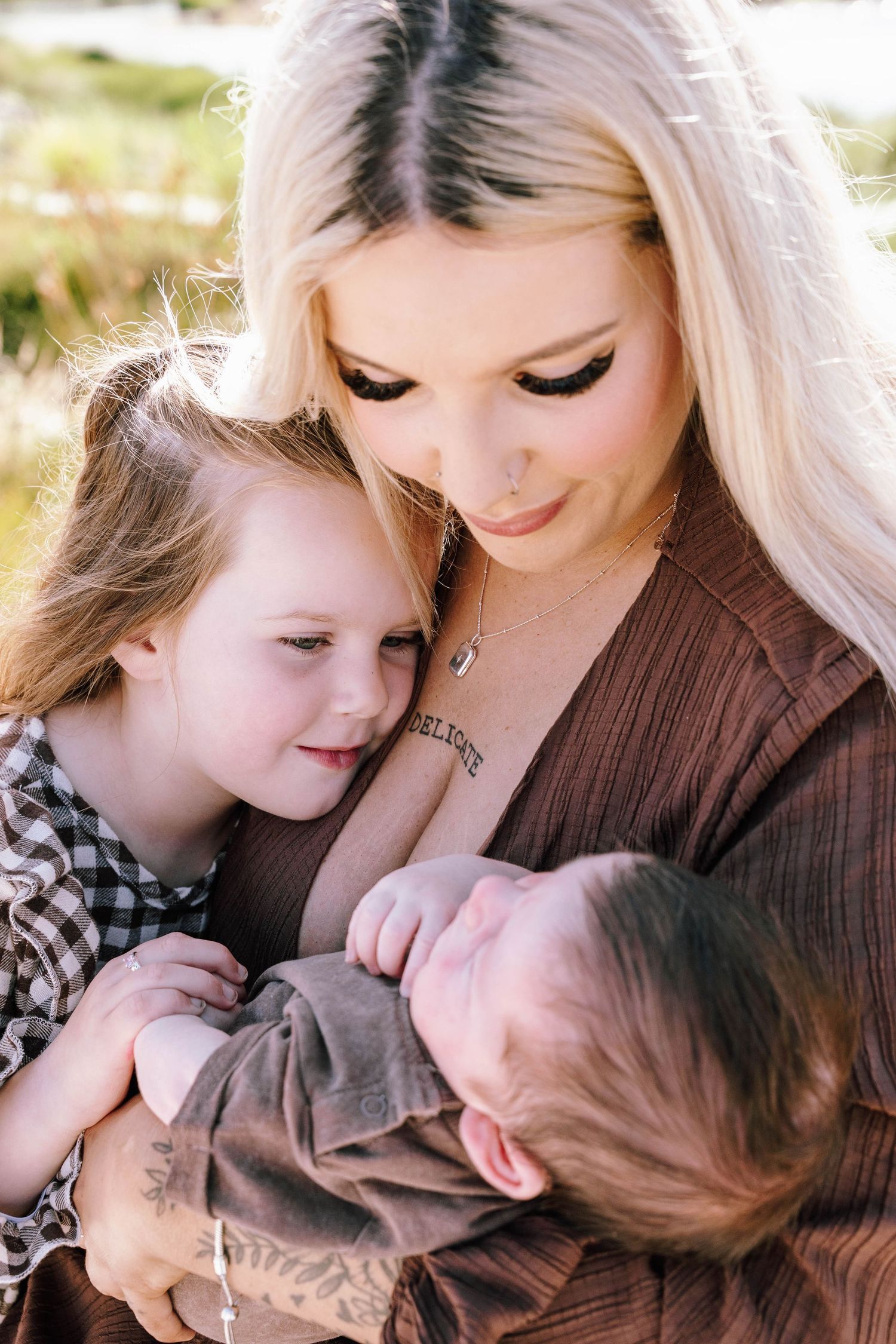 A young blonde mother with tattoos tenderly holds a newborn baby while her toddler daughter leans in close outdoors.