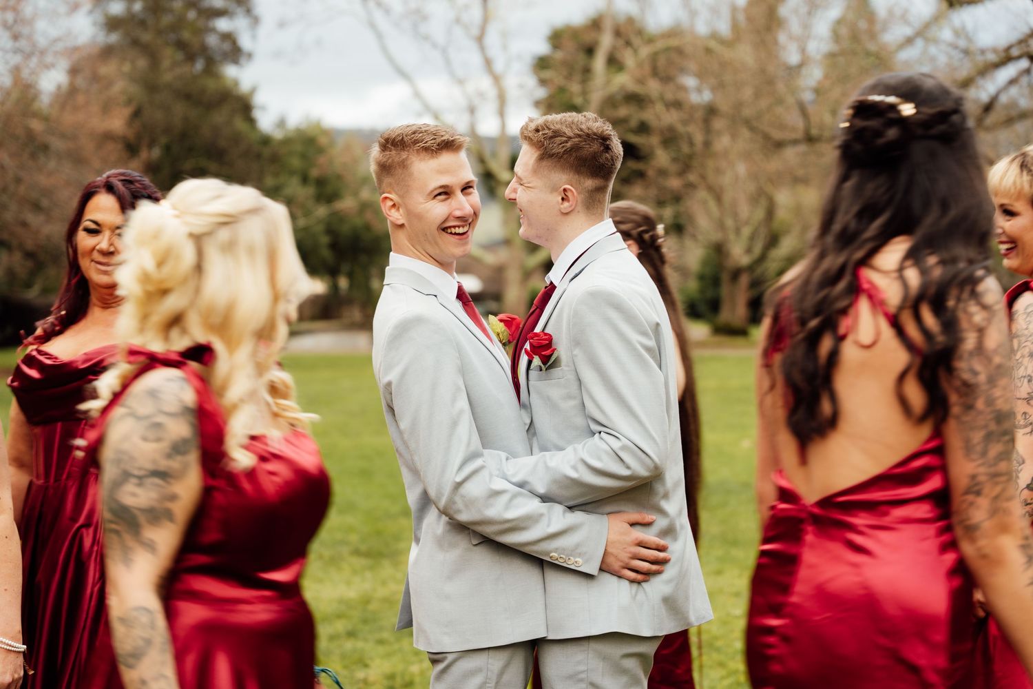 Two grooms in gray suits share a tender moment surrounded by bridesmaids in red dresses at an outdoor wedding.