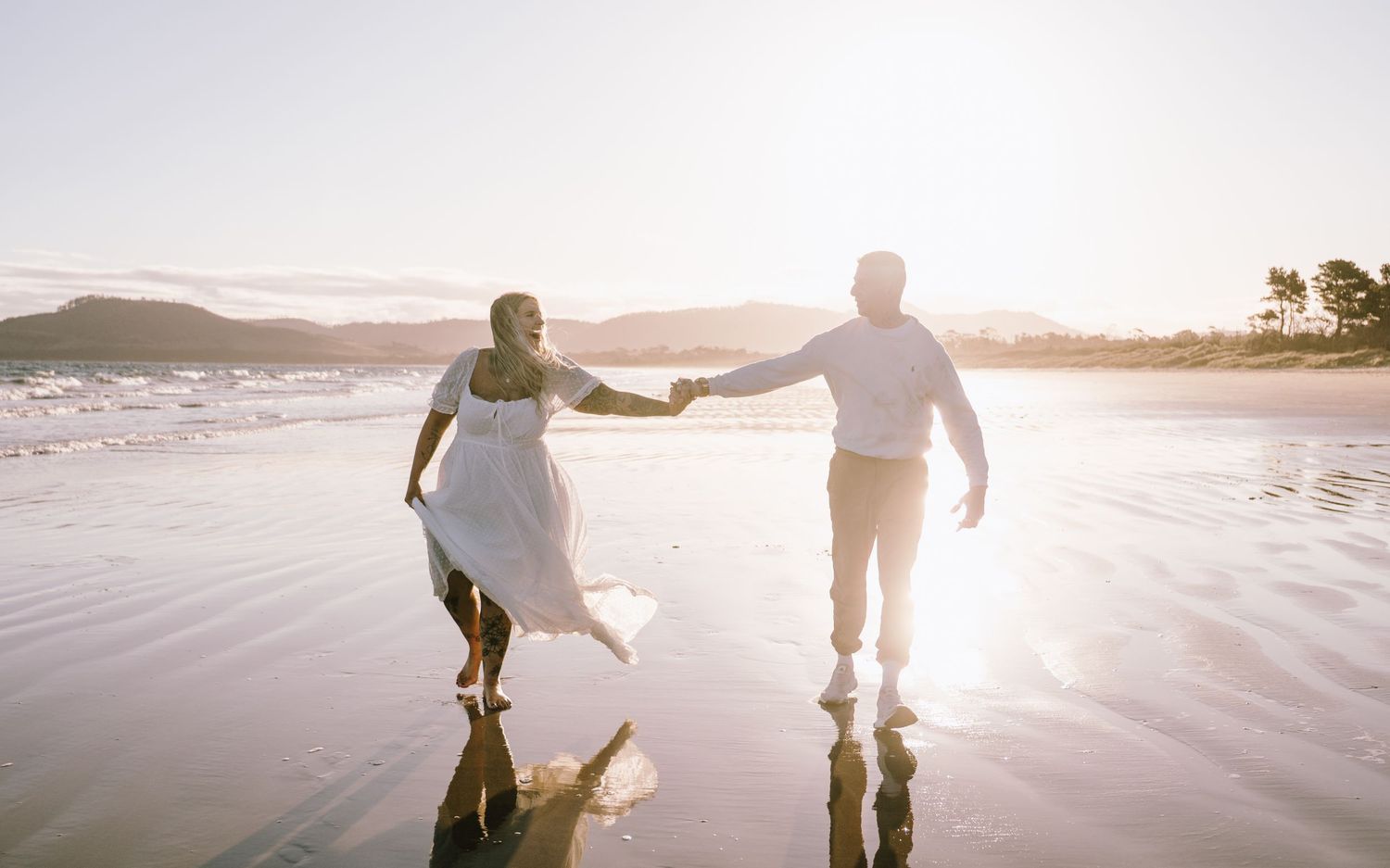 Couple walking hand in hand along a misty beach at sunset with mountains in the background.