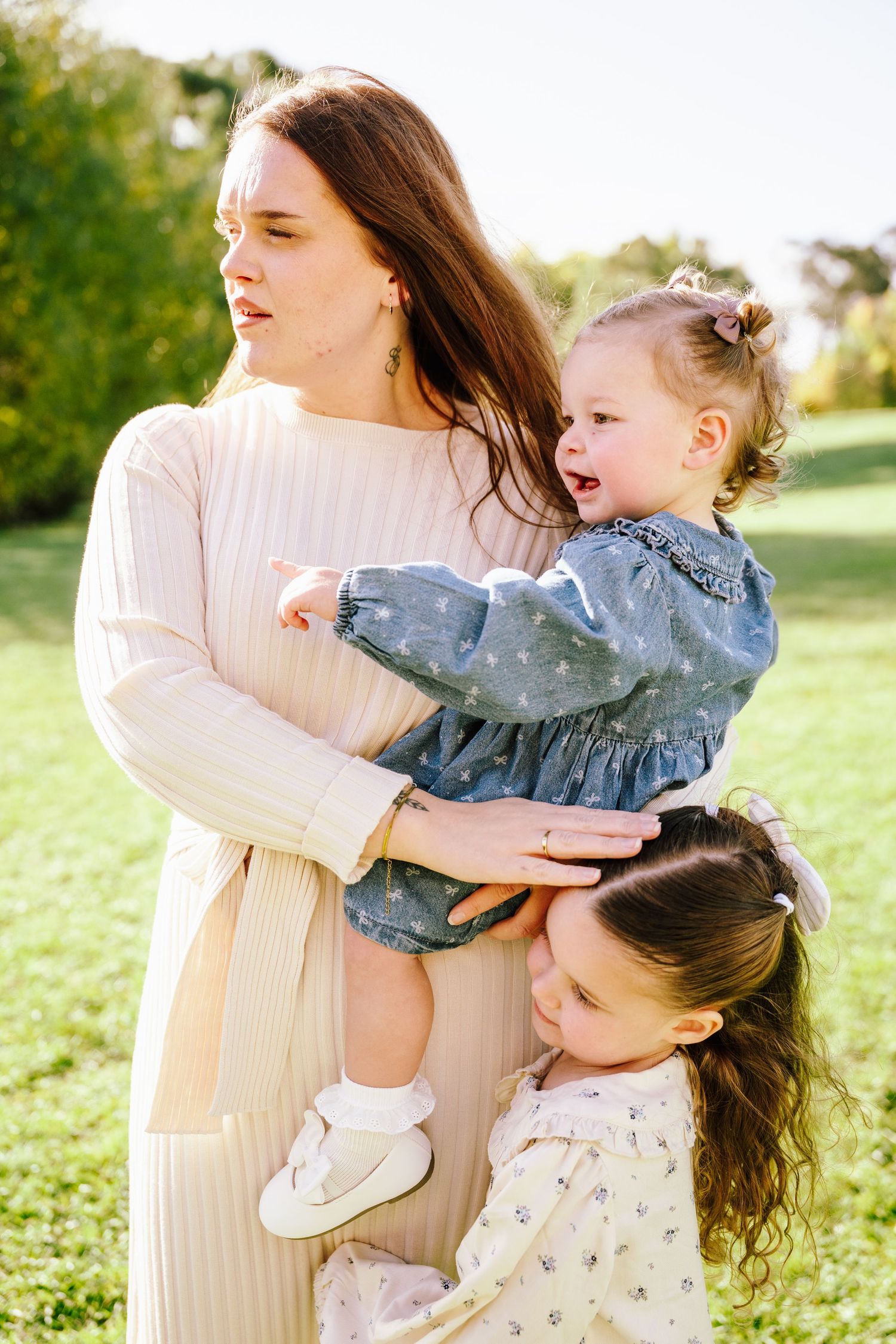 Young mother in beige dress holds toddler while older daughter hugs her in park.