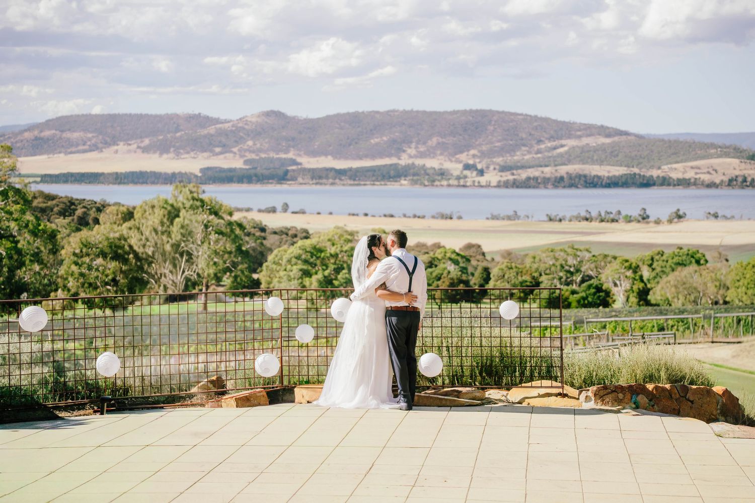 Bride and groom sharing a romantic kiss overlooking a scenic lake and rolling hills at an outdoor vineyard wedding.
