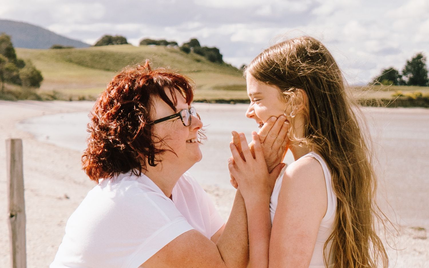 A woman and young girl share a joyful moment on a beach, smiling face to face with green hills behind them.