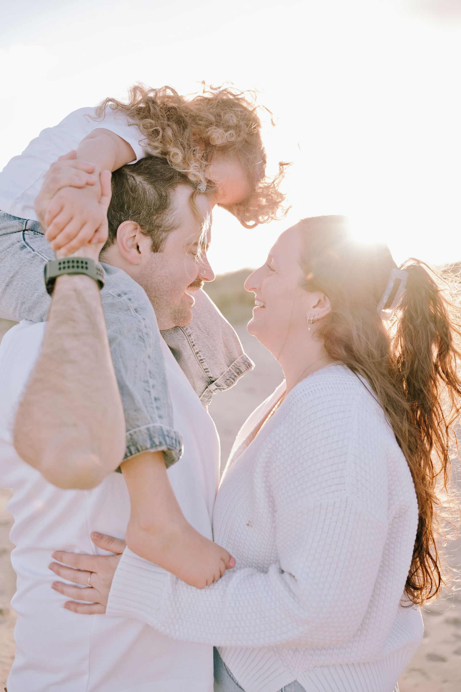 Family moment at sunset on beach, father holding curly-haired child on shoulders while mother smiles joyfully.