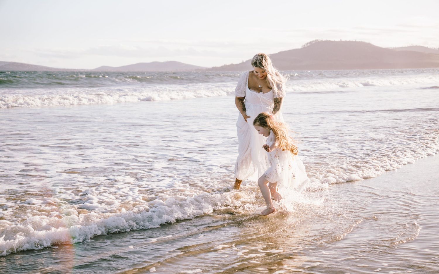Bride in white dress dancing with young girl in ocean waves on sunny beach wedding day.