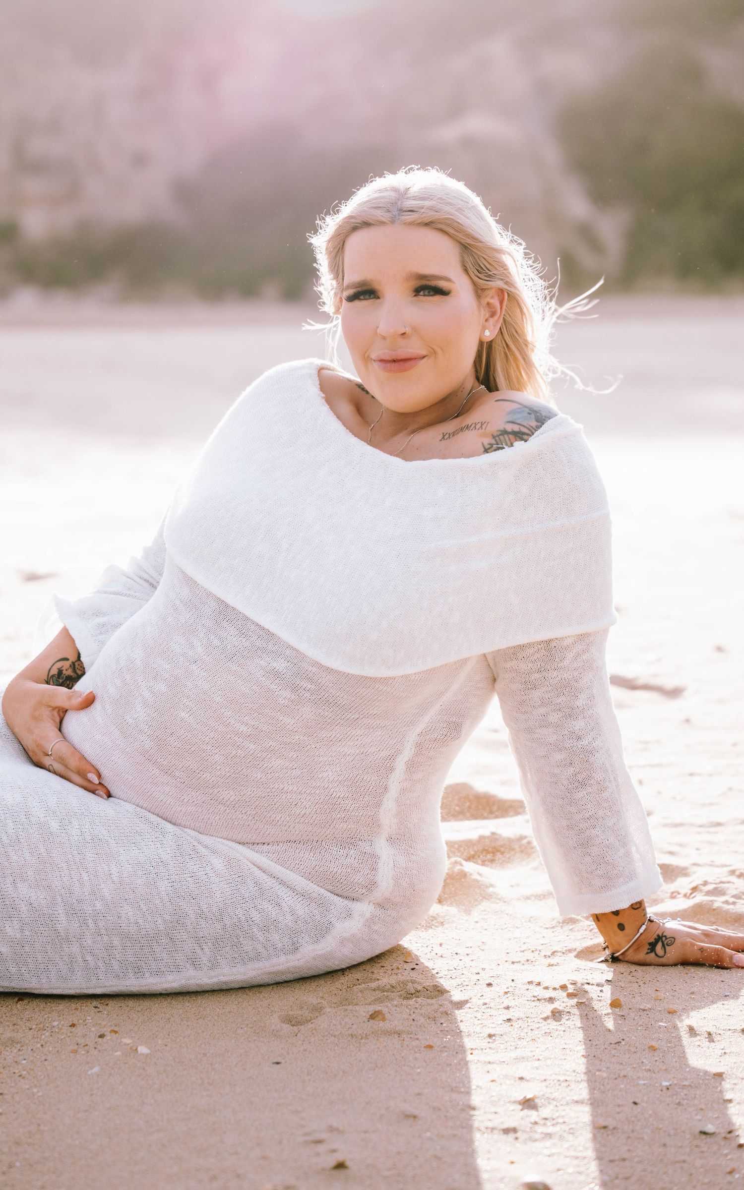 Pregnant woman in white dress sits on beach at sunset with soft lighting and windswept hair.