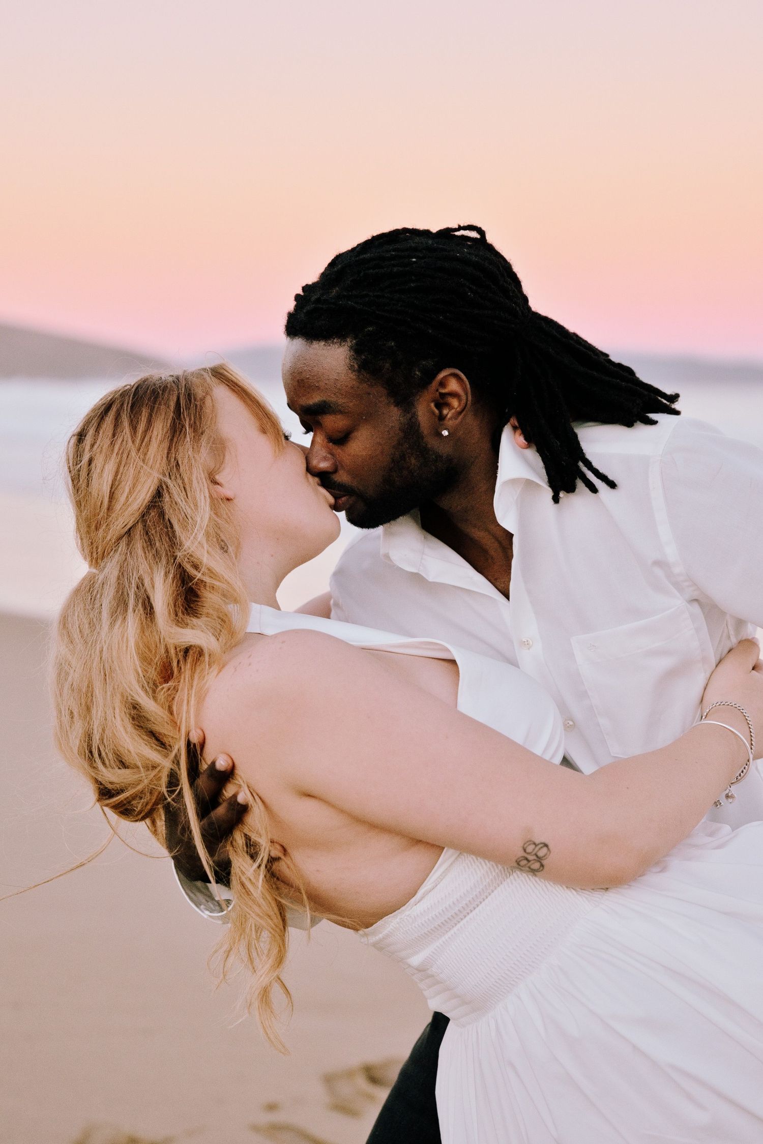 A couple shares a romantic kiss on a beach at sunset, the man in white shirt holding a woman in a white dress.