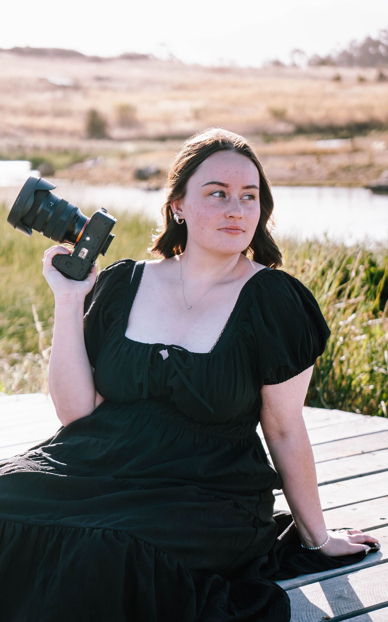 A person in a black dress poses on a wooden bench in a scenic outdoor setting with tall grass and water views.