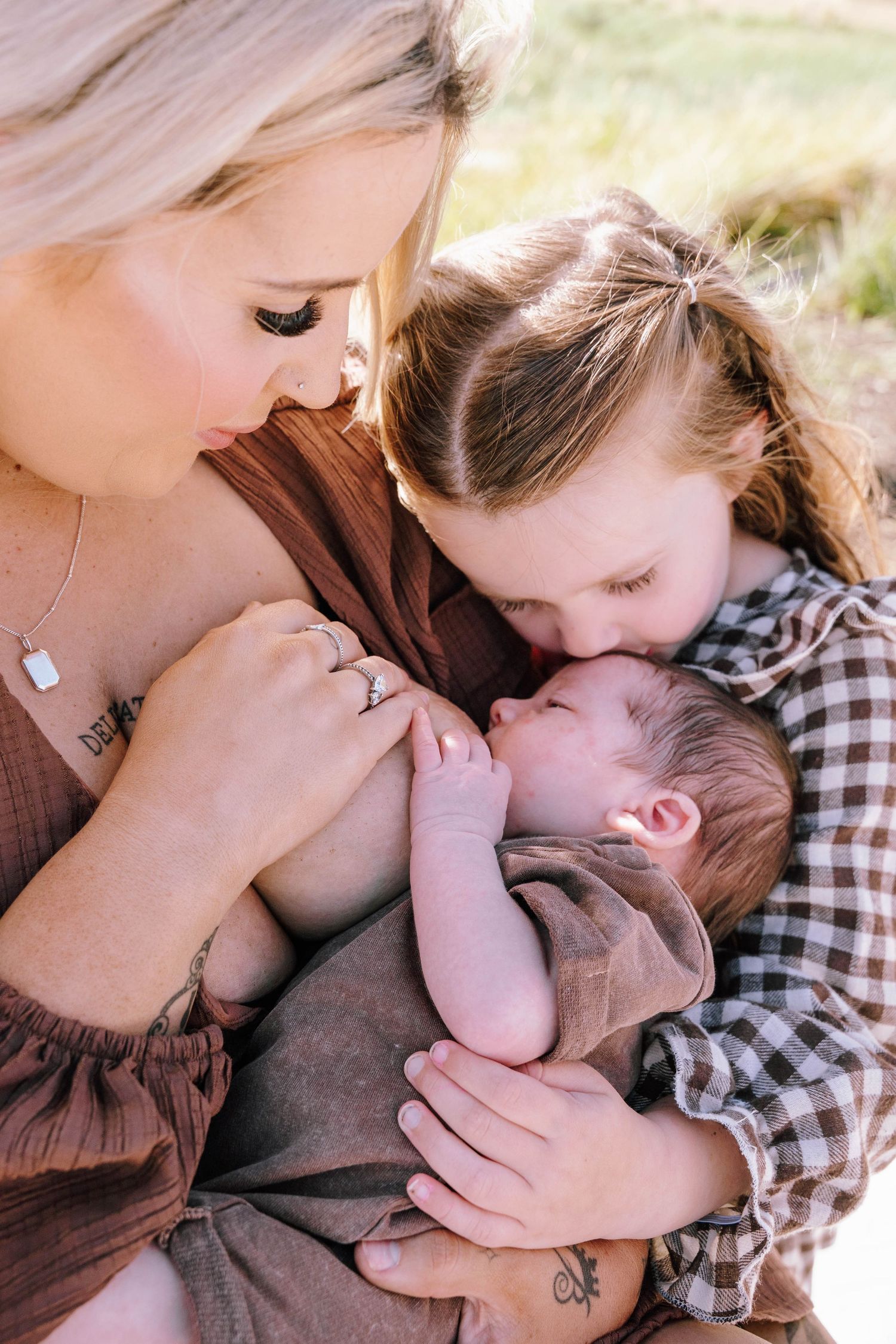 A mother and young child lovingly gaze at a newborn baby outdoors in a warm, tender family portrait.