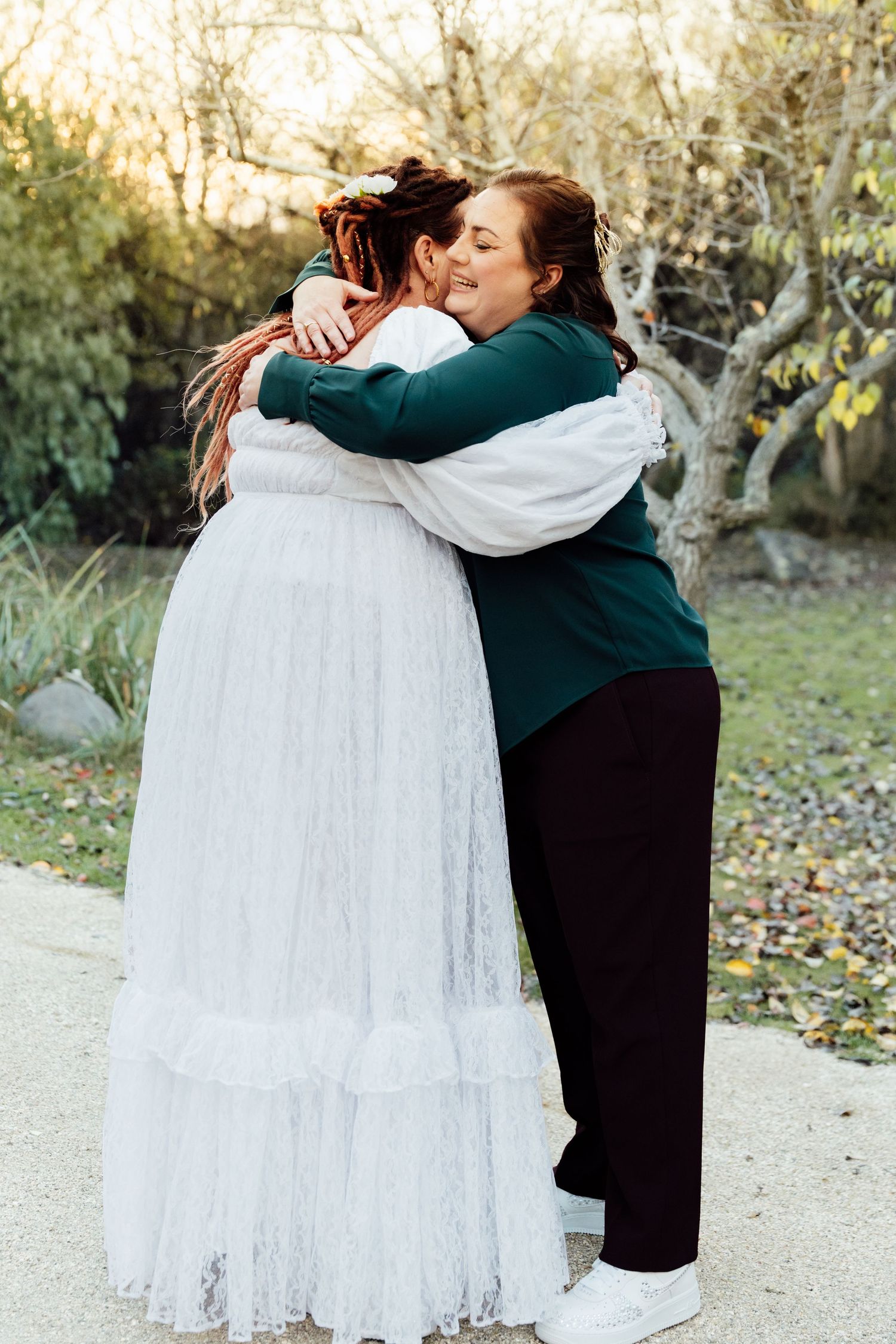Two women share a warm embrace outdoors, one wearing a white lace wedding gown and the other a green sweater.