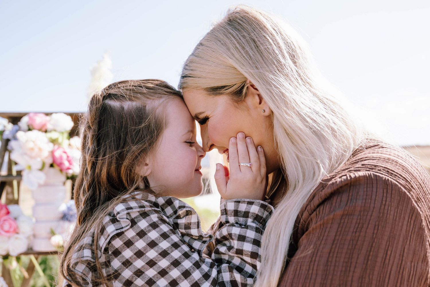 A mother and young daughter share a tender forehead-to-forehead moment outdoors with floral decorations in the background.
