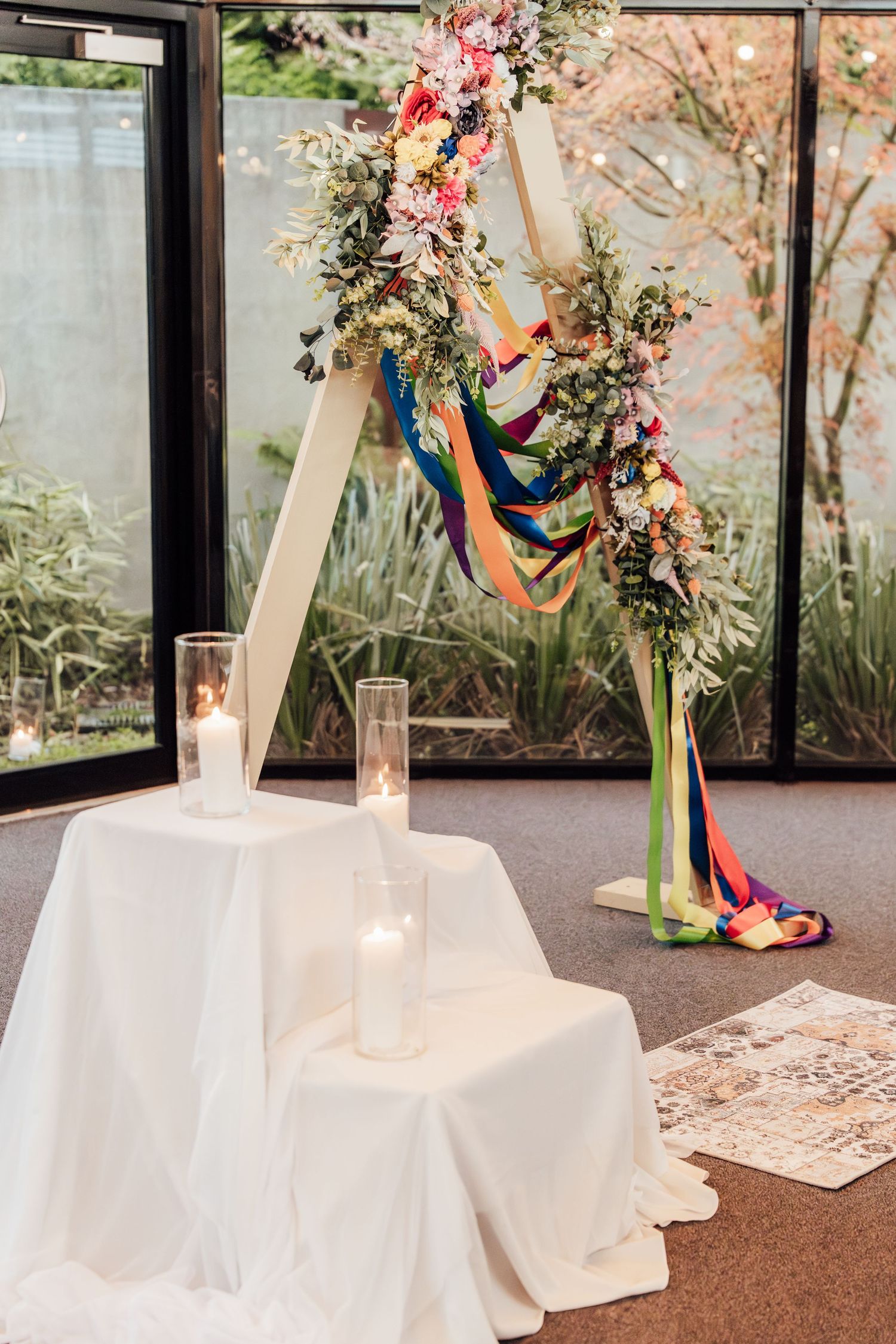 Triangular wedding arch with colorful ribbons and floral arrangements, flanked by candles on white draped tables indoors.