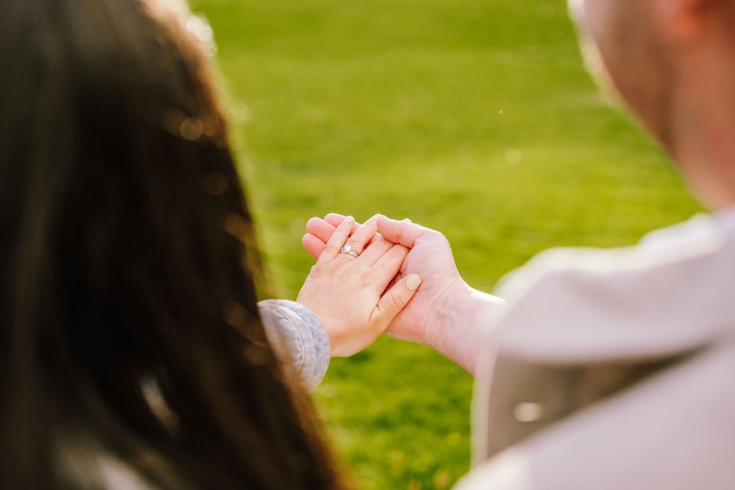 Couple holding hands tenderly outdoors on green grass, woman wearing an engagement ring.